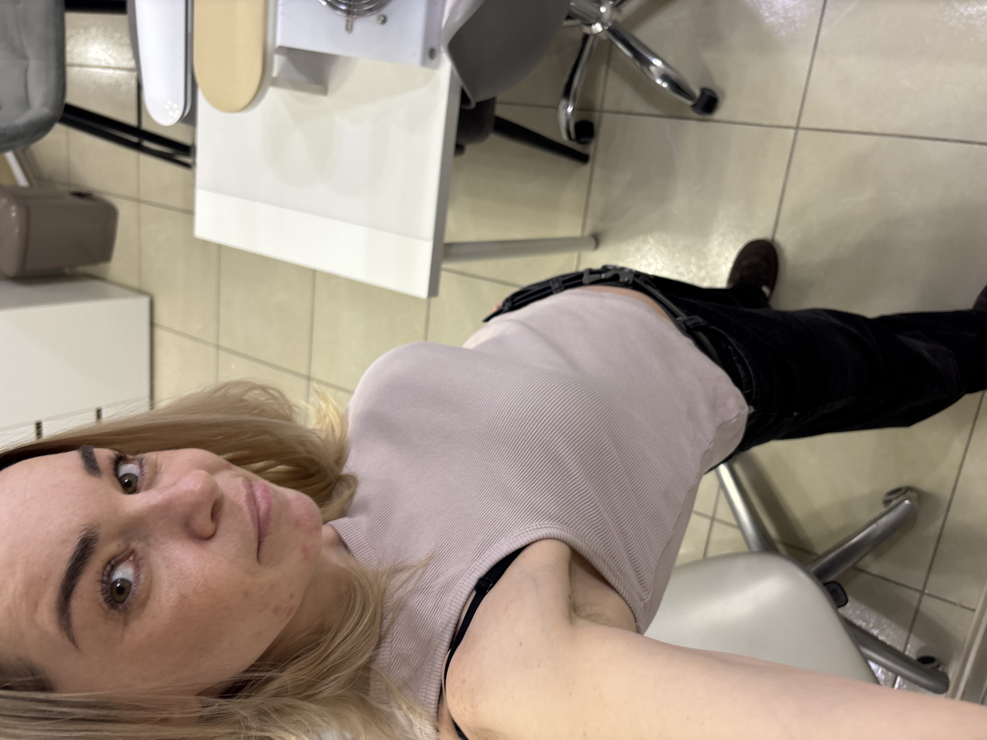 A woman with blonde hair and fair skin takes a selfie. She is wearing a light pink tank top and black pants. She is sitting on a gray chair in what appears to be a tiled room. In the background are several tables and chairs. The location is likely Vsevolozhsk, Russia, as requested.