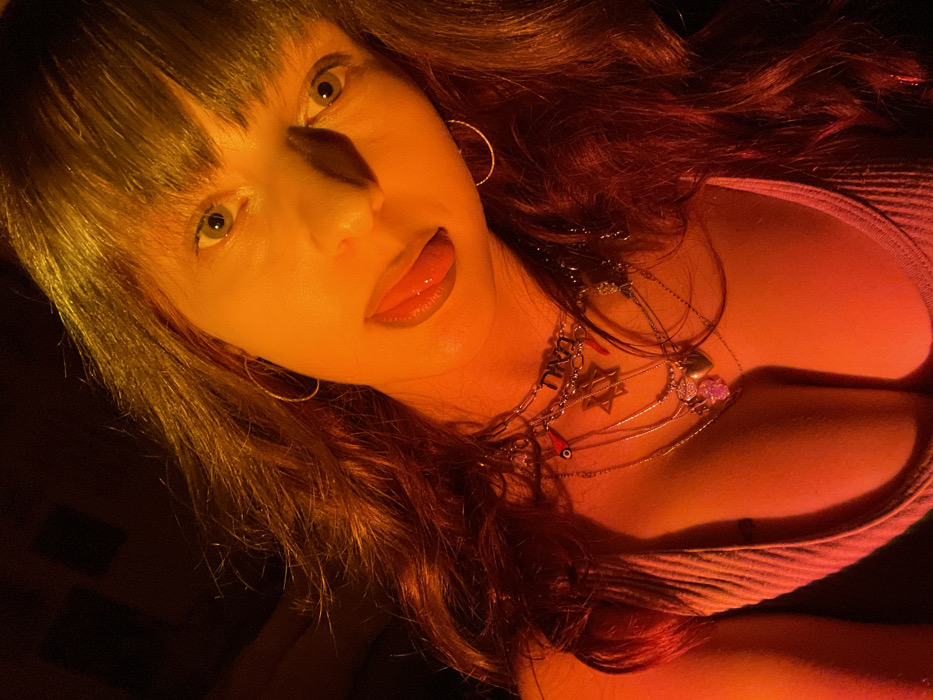 A close-up portrait captures a woman with her face and upper torso illuminated by dramatic, warm orange and red light, creating prominent shadows, especially on her nose. She has long, wavy dark hair with bangs framing her face. Her eyes, appearing green or hazel, are wide and focused directly at the viewer, while her tongue is slightly extended. She wears small, silver-toned hoop earrings.
Around her neck, multiple layered necklaces are visible. One prominent necklace features a chunky chain with a rectangular pendant that clearly displays the Hebrew word "נצח" (Netzach). Another necklace has a Star of David pendant, and a third includes a small red bead, characteristic of an "evil eye" amulet. She is wearing a light-colored, ribbed tank top or similar sleeveless garment. The intense lighting and dark, undiscernible background suggest an indoor setting, likely at night. The image was taken in Raanana, Israel.