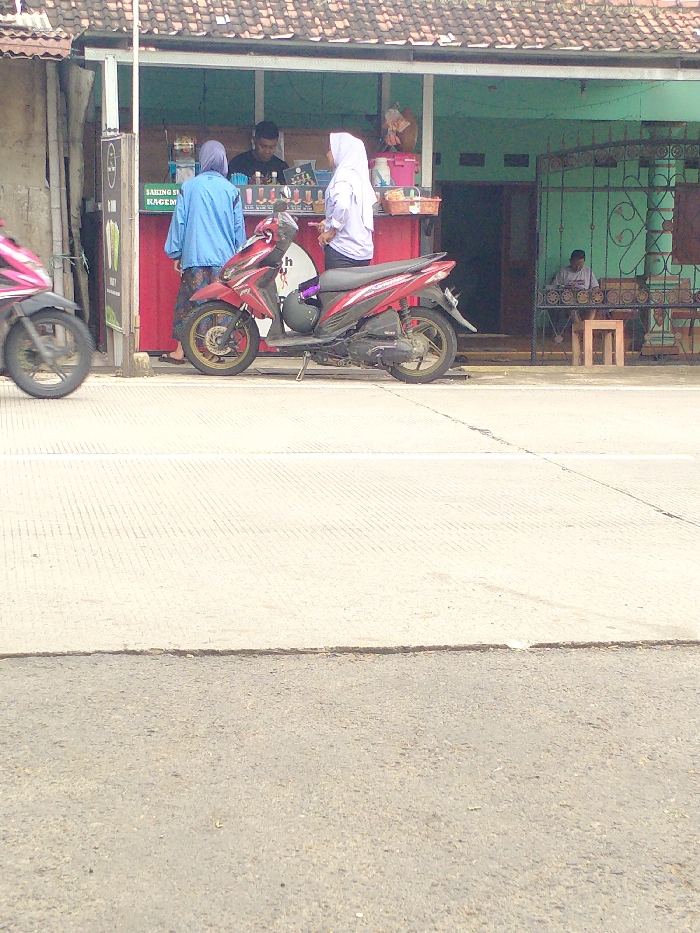 The image captures a vibrant street scene in Kecamatan Kebakkramat, Indonesia, featuring a roadside food stall in front of a building. In the center, two women, both wearing headscarves (hijabs)—one in a blue top and the other in white—are positioned at the red-fronted stall. A male vendor stands behind the counter, attending to them, with various cups and containers of what appear to be beverages displayed.
A red scooter is parked directly in front of the stall, facing right, with a helmet hanging from its handle. On the far left, the front wheel and part of another pink motorbike are visible, suggesting ongoing traffic. To the right of the stall, a man is seated at a wooden table inside a building with a green facade and an ornate metal gate. He appears to be looking down, possibly at a phone or tablet.
The setting suggests a daytime scene with bright, natural lighting and clear weather. The ground in the foreground and midground consists of a light-colored concrete road with horizontal scoring, transitioning to darker asphalt closer to the viewer. On the left side of the stall, a vertical sign is partially visible with text, including "SAKONG SE" and "KAGEM." The overall atmosphere is that of a typical daily commerce activity in an Indonesian urban or suburban area.