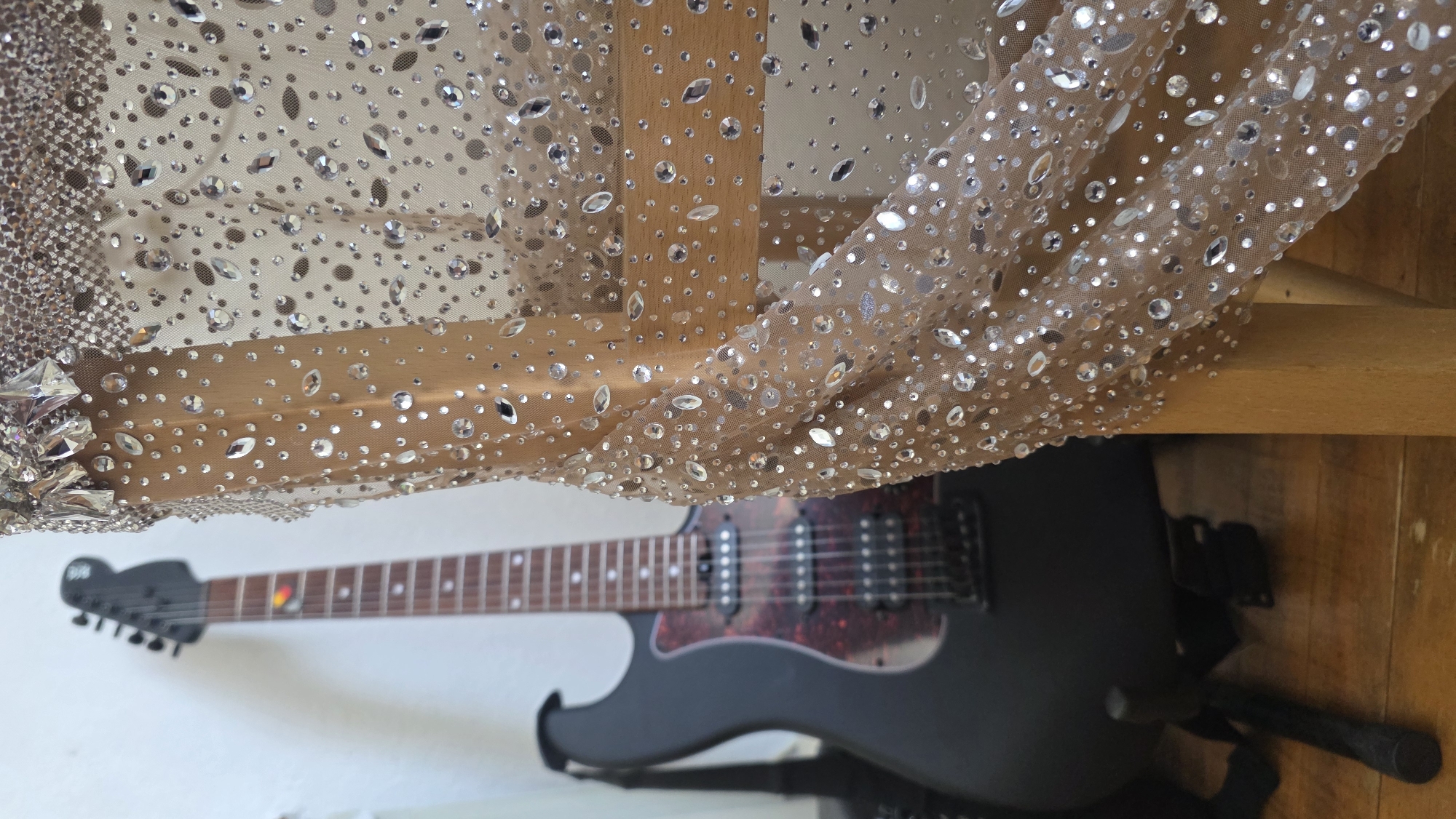 An indoor, medium-shot photograph, likely taken in Blagoevgrad, Bulgaria, captures a glittering, translucent fabric draped over a wooden structure, with an electric guitar positioned below.
The upper and central part of the image is dominated by a sheer, light beige or nude-toned fabric, possibly mesh or tulle, which is profusely embellished with numerous sparkling rhinestones and crystals. These rhinestones vary in size and shape, including small circular gems, larger round crystals, and elongated oval or marquise-cut stones, all reflecting light brightly. A section on the far left of the fabric features a denser, more intricate pattern of smaller crystals and larger faceted gems. The fabric is draped in soft folds, revealing a light wooden beam or frame underneath.
Below the shimmering fabric, a matte black electric guitar is visible, oriented horizontally. It features a dark fretboard with light dot inlays, a distinctive red and yellow inlay around the third fret, and a dark headstock with an indiscernible white logo. The guitar's body has a red and black tortoiseshell pickguard and two black humbucker pickups. It appears to be resting on a black stand.
The background consists of a light-colored wall on the left and a wooden surface, possibly a floor or another piece of furniture, on the right. The lighting suggests an indoor setting, likely during the day, with ample light causing the rhinestones to glint. There is no explicit activity taking place; the image presents a still life arrangement focusing on the contrasting textures and materials. No discernible text is clearly visible other than the faint logo on the guitar headstock.