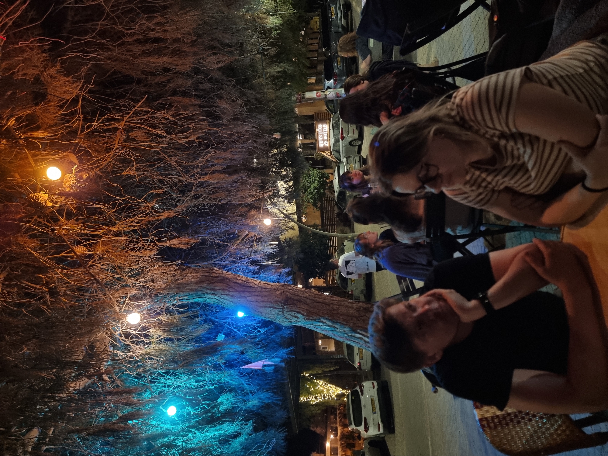 This image depicts a nighttime scene in Mitzpe Ramon, Israel. A group of people are seated at tables, likely at an outdoor restaurant or bar. The foreground features the top portion of a man with short, dark hair, wearing a black shirt and a watch. He is leaning on his hand. Another woman with glasses and a striped shirt is visible next to him. Further back, more people are seated, some looking up. The background contains a large tree illuminated with orange and blue lights. Cars are parked in the street behind the seating area. A sign is visible, but the text is not discernible. String lights illuminate a building further back.