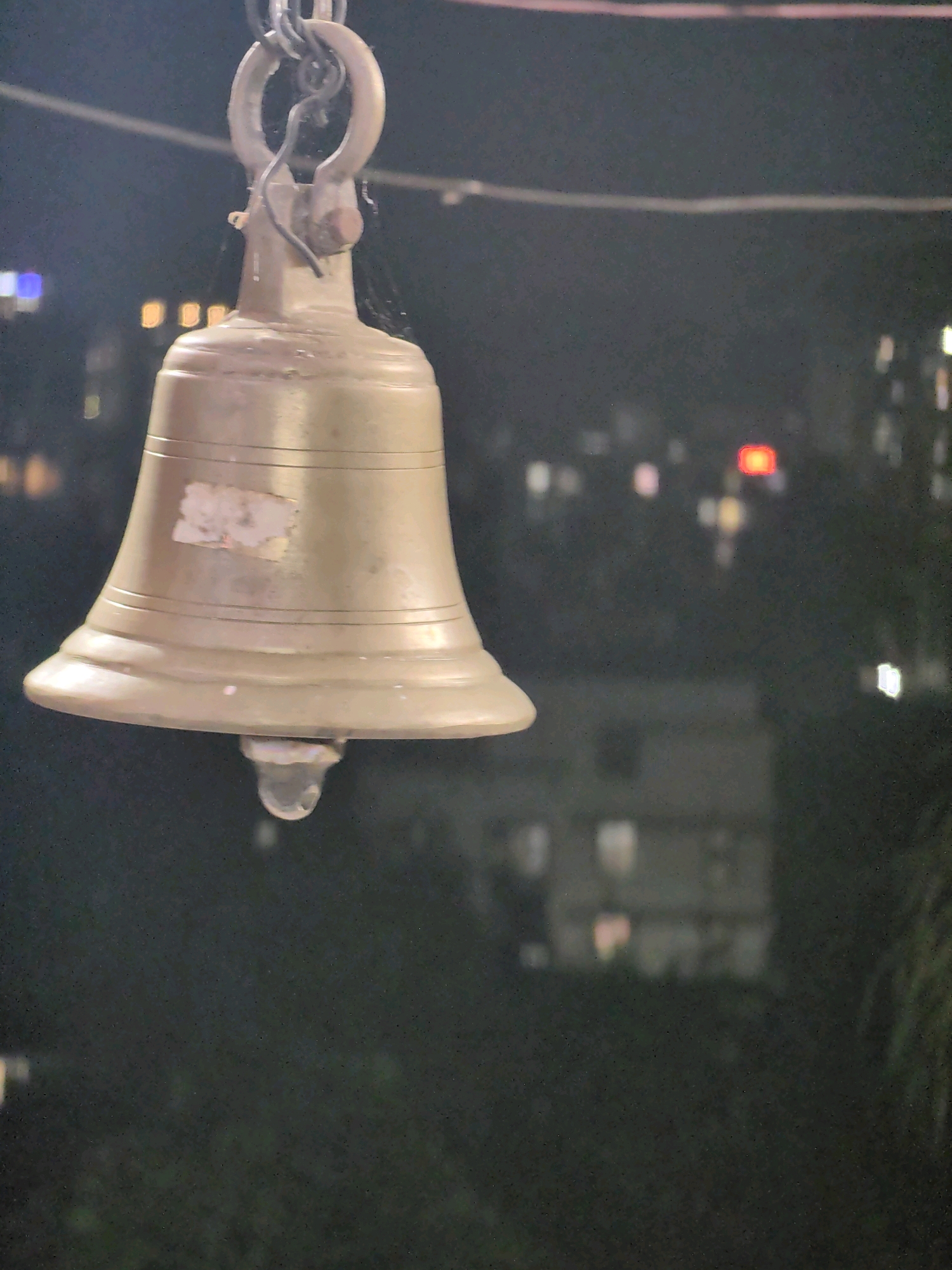 A close-up, slightly blurred, eye-level shot features a golden bell hanging from a chain against a dark, out-of-focus background depicting a city at night, likely Mumbai, India. The bell is the primary subject, suspended centrally and dominating the foreground. It has a classic bell shape with distinct ridges and a small clapper hanging from its center. A small, unidentifiable sticker is affixed to the side of the bell. The background shows a soft bokeh of city lights, suggesting buildings and a general urban environment. The lighting is dim, with the bell being the most illuminated object. No people are visible in the frame. The overall mood is calm and possibly serene, with the focus on the solitary bell.