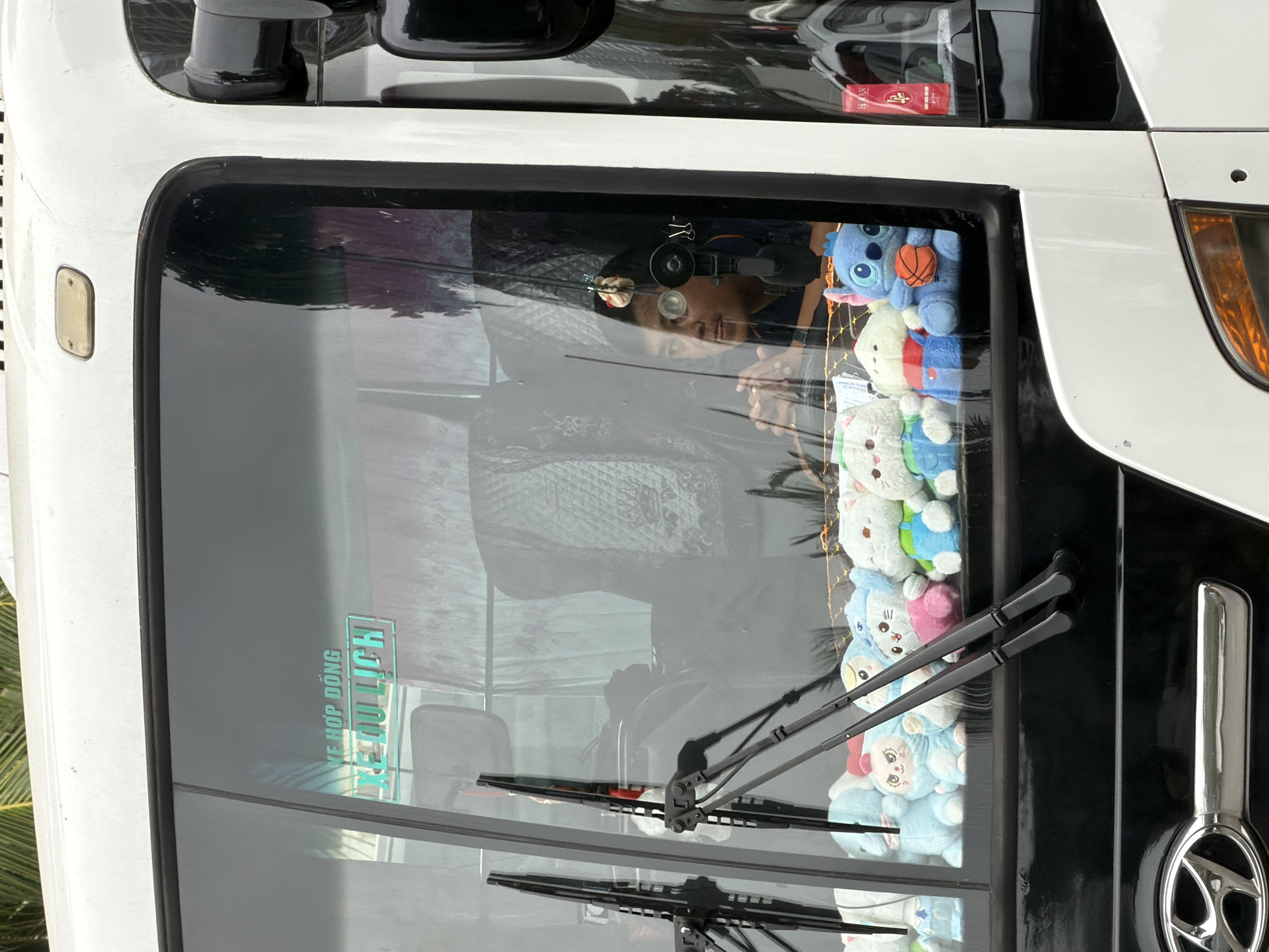 Captured at a slightly canted angle, the close-up image features the front window of a white bus, likely a passenger vehicle, reflecting the seated driver and an array of stuffed animals lining the inside of the windshield. The windshield also bears the words "XE HOP DONG XE DU LICH," potentially indicating a tourist transport or charter bus service. The driver, a dark-haired person, is visible through the glass, with their hands clasped. Two sets of windshield wipers are present, indicating functionality. The surrounding environment suggests the bus is likely located in the city of Đà Nẵng, Vietnam, based on the language used on the windshield.
