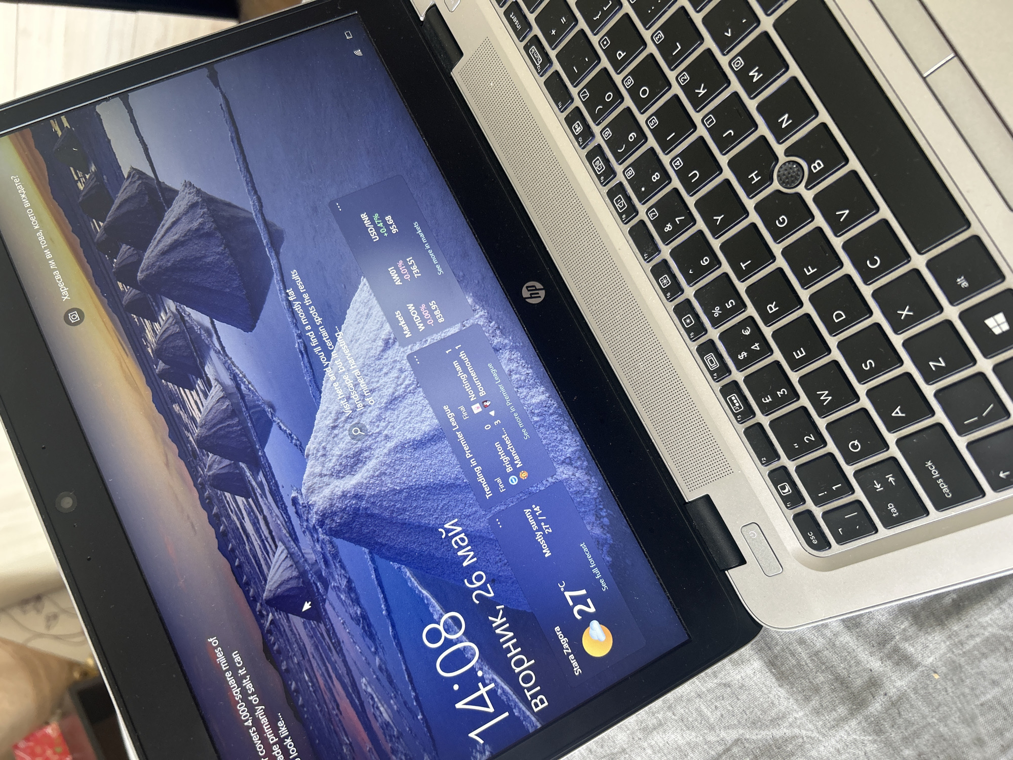 The image shows a laptop, partially open, displaying a Windows lock screen. The time displayed is 14:08 on Tuesday, May 26th. The location is listed as Stara Zagora, with a current temperature of 27°C and a forecast of mostly sunny conditions, with a low of 14°C. Weather forecasts, trending information, and market data are displayed.