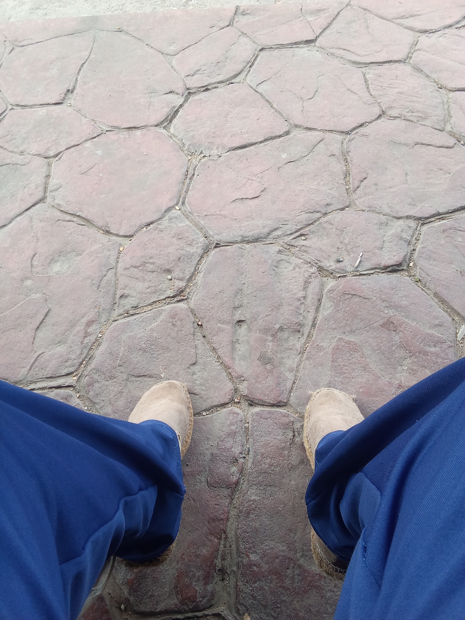The image shows a downward-facing perspective of a person sitting on a red-brown, irregularly shaped stone pavement. The person is wearing blue pants and light beige espadrille shoes. The background is the pavement made of stones and with a small amount of loose dirt. The location cannot be determined from the given image.