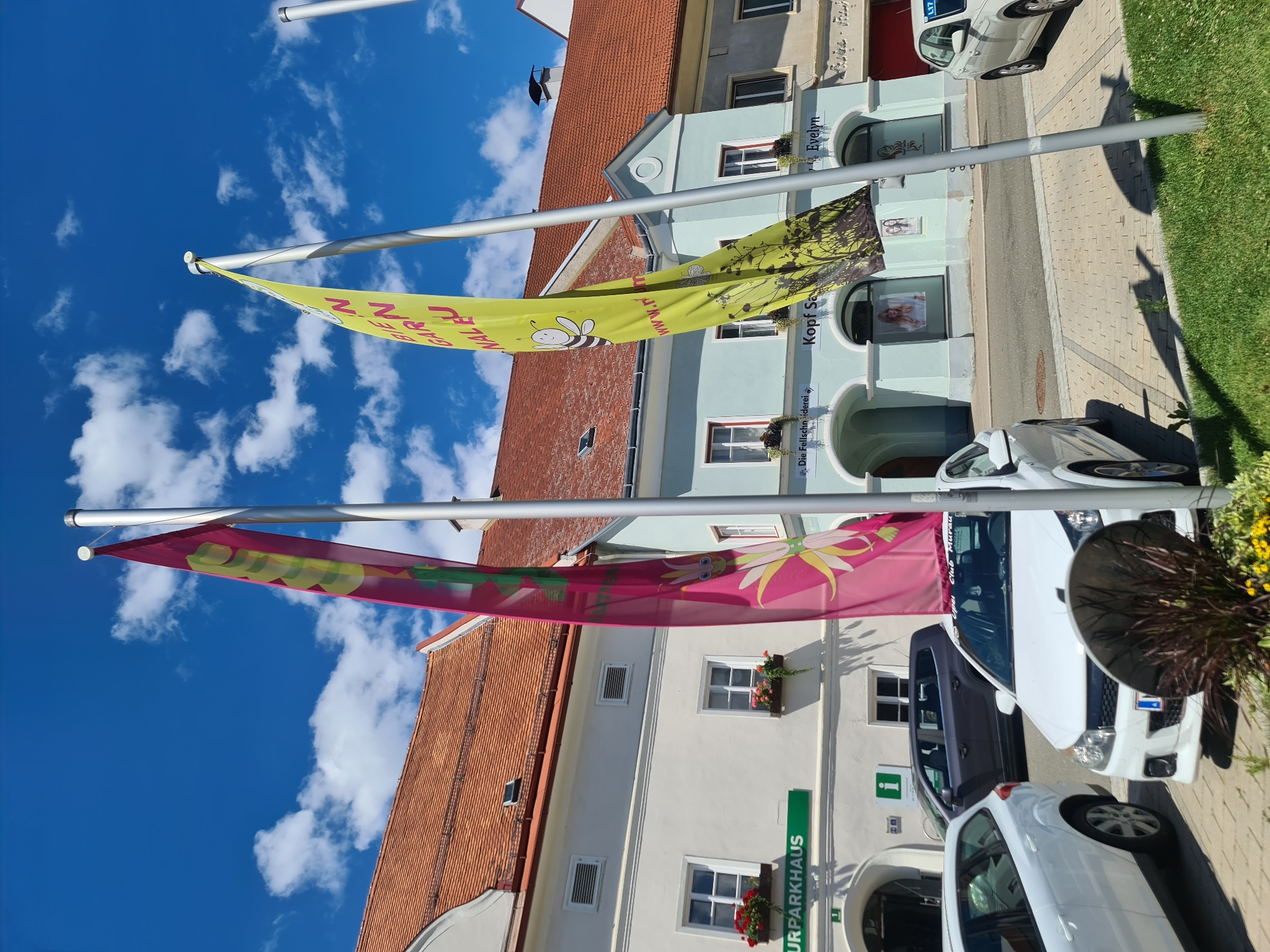 A low-angle, vertical shot captures two vibrantly colored flags flying from poles against a brilliant blue sky with fluffy white clouds. The flags are oriented horizontally, with the top one being yellow and adorned with a cartoon bee and leafy designs, along with partial text that appears to read "GARLZ NAL F www.r". The bottom flag is a bright pink and features a stylized yellow flower with a cartoon face.
Behind the flags, a street scene unfolds. Buildings with red tile roofs and light blue or white facades line a cobblestone street. Storefronts are visible, with one featuring the text "Evelyn" and another "Kopf Sa". Further down, the text "Die Fellschn siderei" is also visible. The street is quiet, with a few white cars parked along the side. One car has the partial text "Club Murau" visible on its windshield. Signs for "URPARKHAUS" are also present.
The scene suggests a sunny day in a European town. The flags might be part of a local festival or event, adding a festive touch to the otherwise ordinary street. The bright colors of the flags and the clear blue sky evoke a sense of cheerfulness and a pleasant atmosphere. The overall impression is one of a charming, well-maintained town on a beautiful day.