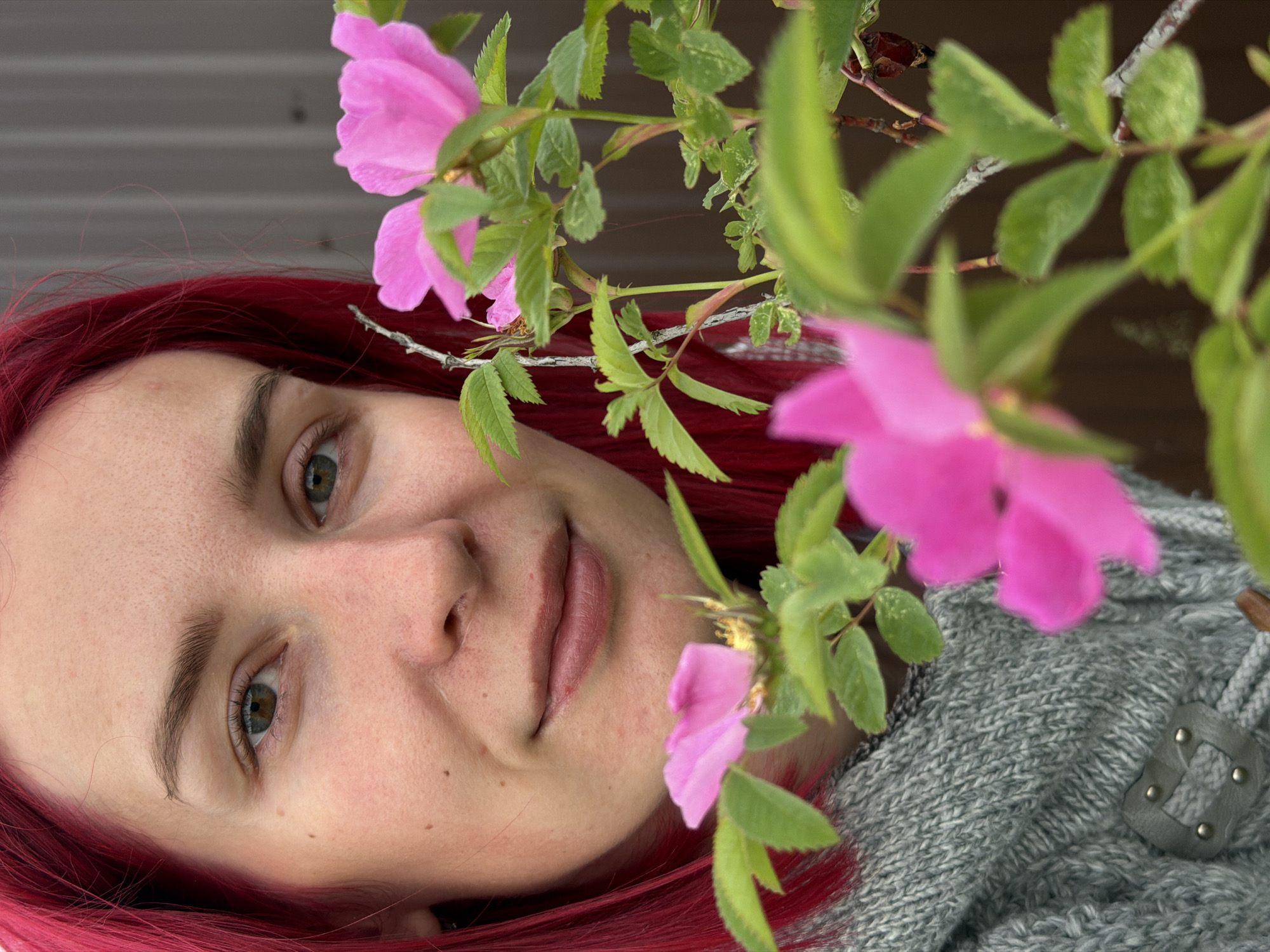 A woman with red hair lies facing up next to pink flowers. She is wearing a grey sweater. The photo was taken in Chaikovsky, Russia.