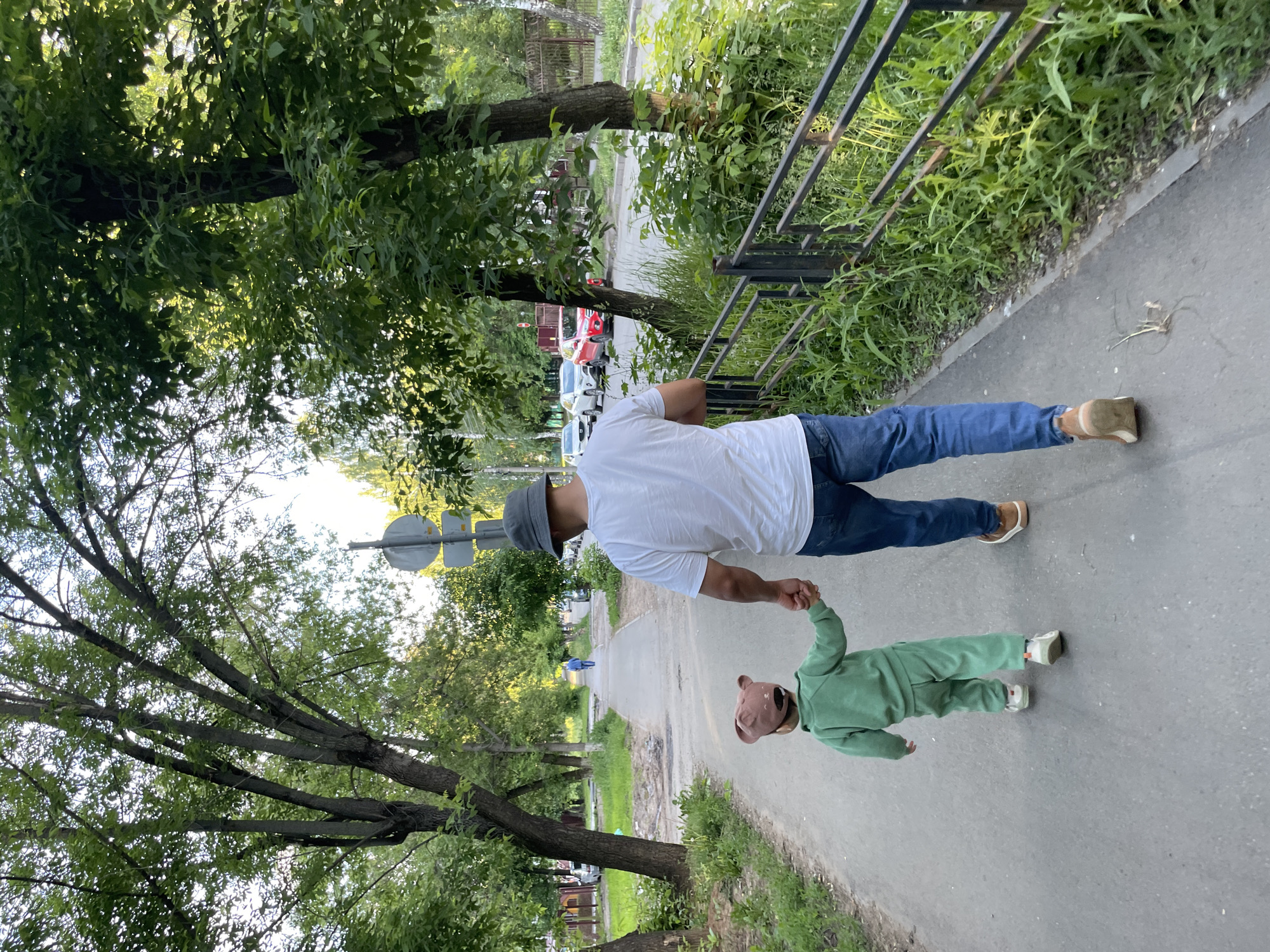 An adult male and a small child are captured walking hand-in-hand on a paved pathway, likely in a park or tree-lined street in Izhevsk, Russia. The adult, viewed from behind, is dressed in a white t-shirt, blue jeans, brown shoes, and a grey bucket hat. The child, also seen from behind, wears a green tracksuit and a distinctive brown hat with ear-like protrusions. They appear to be strolling casually, with the adult holding the child's right hand.
The scene is set outdoors during bright daylight, suggesting a warm season like spring or summer. Lush green trees with dense foliage line both sides of the pathway, creating a shaded and verdant environment. To the right, a black metal fence runs alongside the path, bordering a grassy area. In the background, beyond the trees, a street with several parked cars is visible, indicating a residential or semi-urban area. A round road sign is partially obscured by trees in the middle distance. Another person can be faintly seen walking further down the path. The overall atmosphere is one of calm and leisure. No visible text is present in the image.
