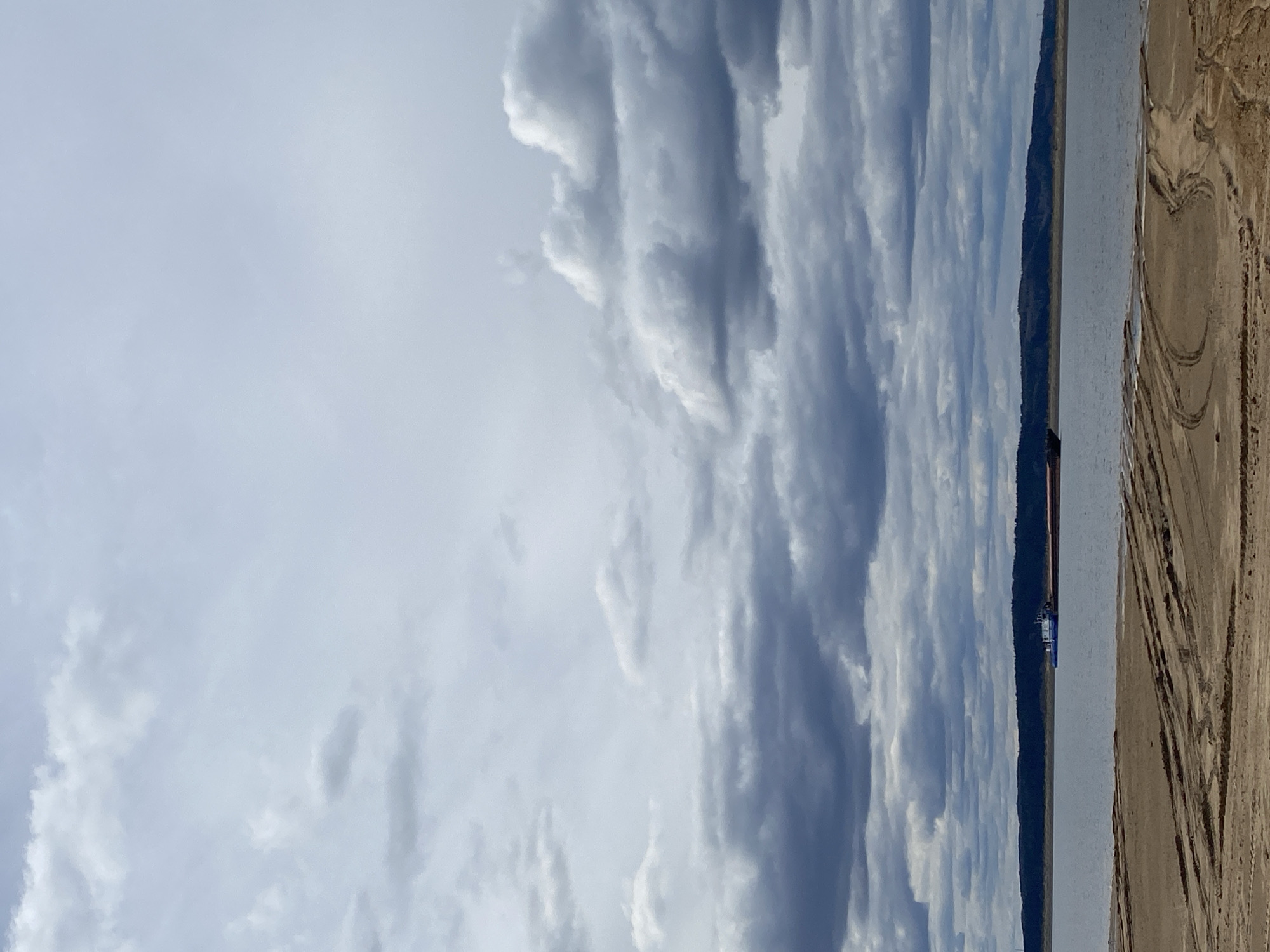The image shows a landscape scene in Chaikovsky, Russia. The sky is mostly covered with striated clouds. Below the clouds is a flat body of water. On the right side of the image is a sandy or earthen area with tire tracks. A distant landmass is on the far side of the water. A boat is partially visible on the water.
