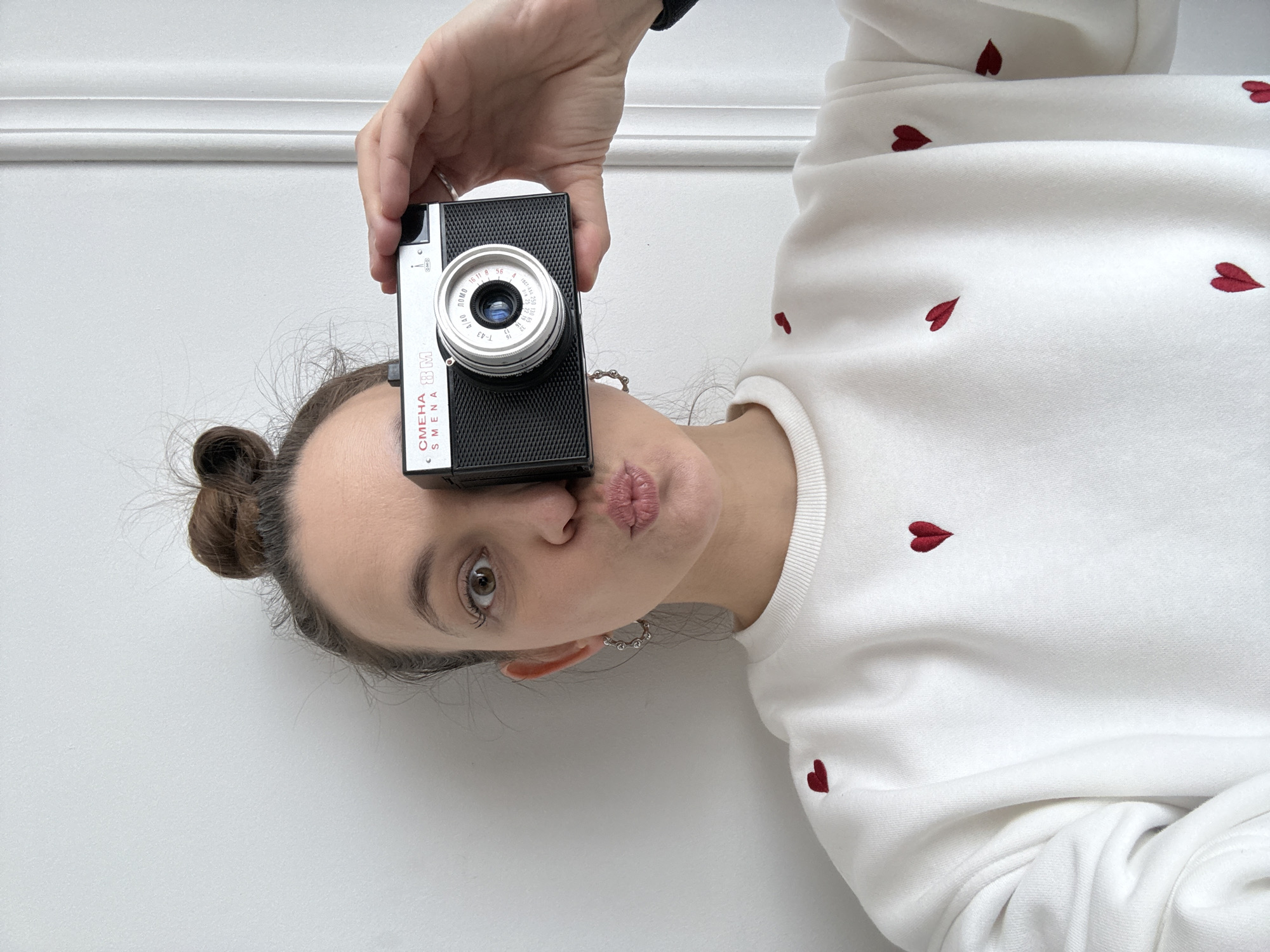 A woman is holding a Смена (SMENA) 8M camera up to her face, making a kissing expression. The camera, labeled with "ЛОМО" (LOMO) and various aperture settings in Russian and English, is positioned in front of her face. She is wearing a white sweatshirt adorned with small red heart embroidery and silver hoop earrings. Her hair is styled in a bun. The background features a plain white wall with a decorative molding near the top. The photograph is oriented sideways.