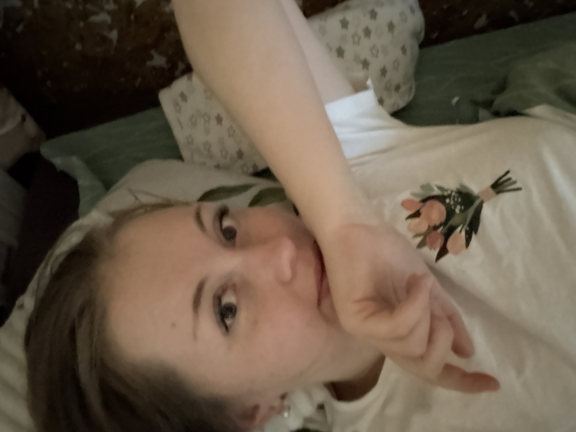 The image shows a woman lying on a bed, with her hand near her mouth. She is wearing a white shirt with a floral design. The bed has green sheets and a pillow with a star pattern. The location is Unknown, Unknown.
