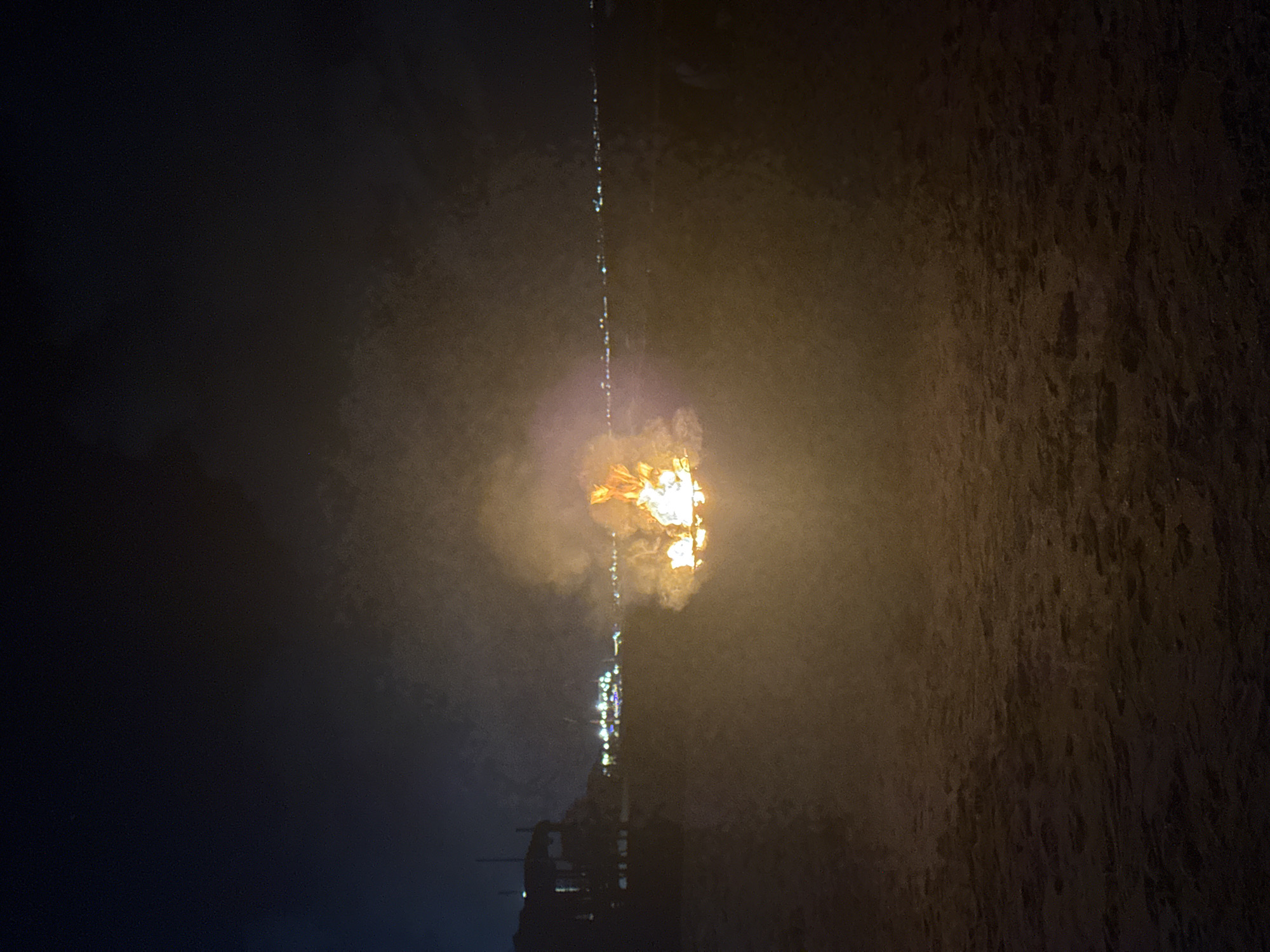 The image shows the Eternal Flame monument in North Kuta, Indonesia, at night. The flame, a bright orange-yellow, burns atop a dark structure, possibly a tower. A thin streak of light, possibly water droplets, extends upwards from the flame. The surroundings are obscured by darkness and fog or smoke, with a textured dark area on the right possibly representing a wall or building. The lower portion of the structure holding the flame is visible.