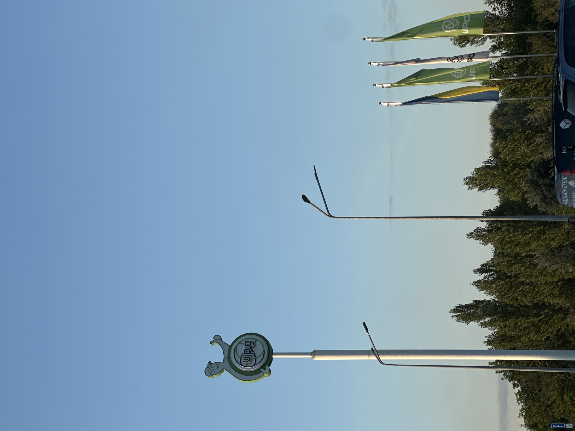 The image shows a gas station sign with a cartoon character and the "UPG" logo. There are flags flying in the background, including the Ukrainian flag. The scene appears to be an outdoor setting, possibly a parking lot or roadside, with trees and a clear blue sky. The UPG gas station is located in Kamianets-Podilskyi, Ukraine.