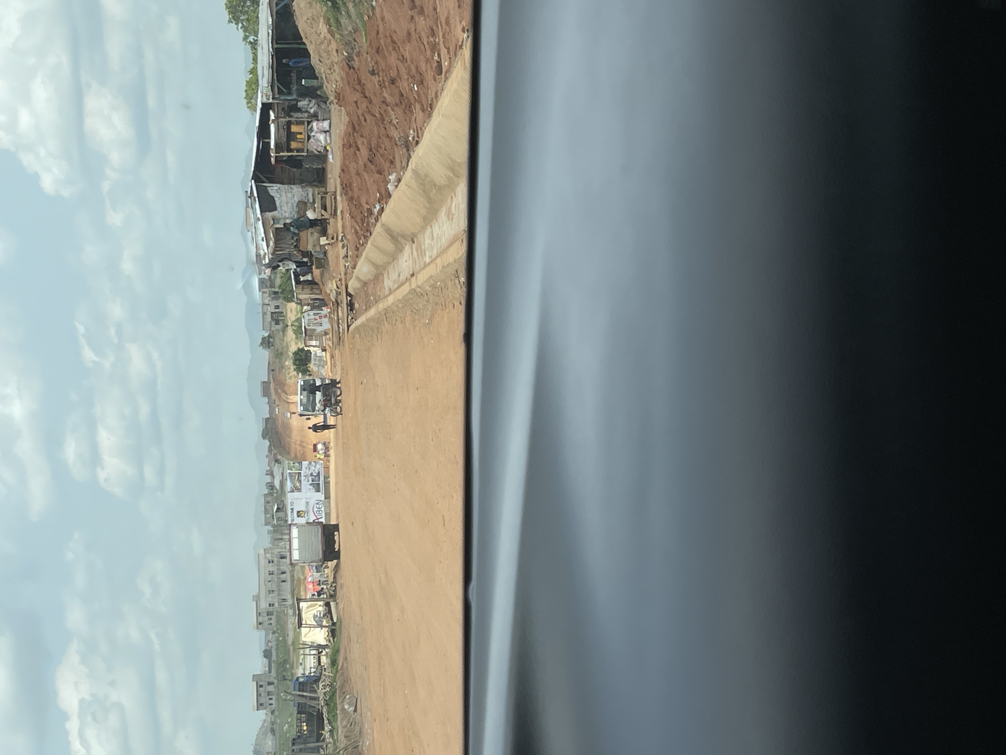 The image shows a view of a dusty, unpaved road in what appears to be Angwan Sayawa, Nigeria. The scene is captured from inside a vehicle, with a portion of the vehicle's interior visible on the right side of the frame. Along the left side of the road, there are makeshift structures and buildings of varying sizes, indicating a bustling commercial or residential area. A sign is visible, partially obscured, appearing to read "Welcome to." Several vehicles and motorcycles are present, along with people walking along the road. The sky is overcast with clouds. The landscape is somewhat uneven, with indications of construction and undeveloped areas along the edges of the road.