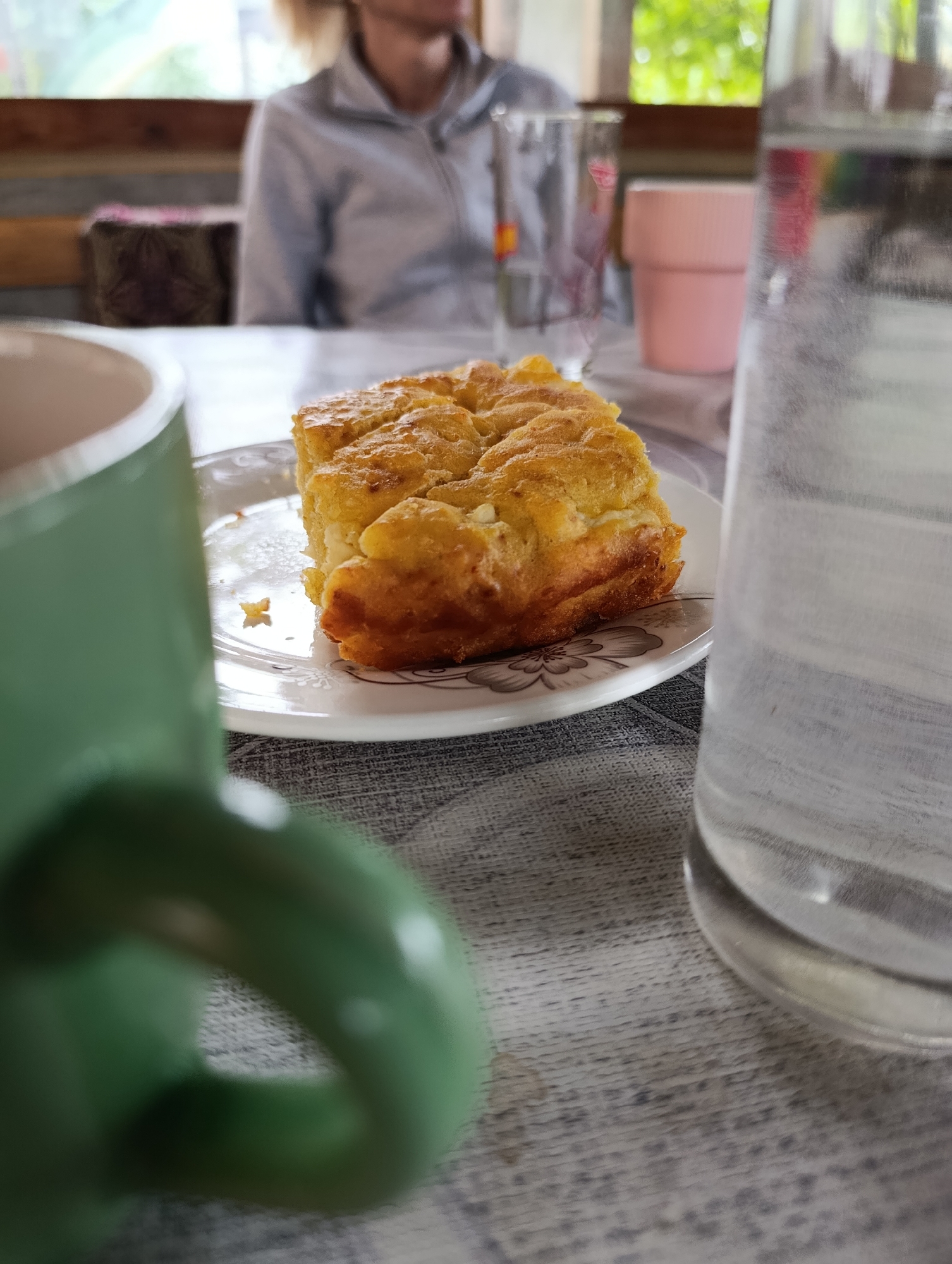 In Mladinovo, Bulgaria, a close-up shot captures a slice of golden-brown baked goods on a white plate with a floral pattern. The pastry appears to be a type of cake or pie, with a slightly uneven, textured surface. To the left, a blurred green mug is in the foreground. To the right, a glass of water is partially visible, its surface showing condensation. In the background, a person wearing a light-colored zippered jacket is seated at a table, their face and hair out of focus. Behind them, a window offers a glimpse of greenery. The table is covered with a gray textured cloth.