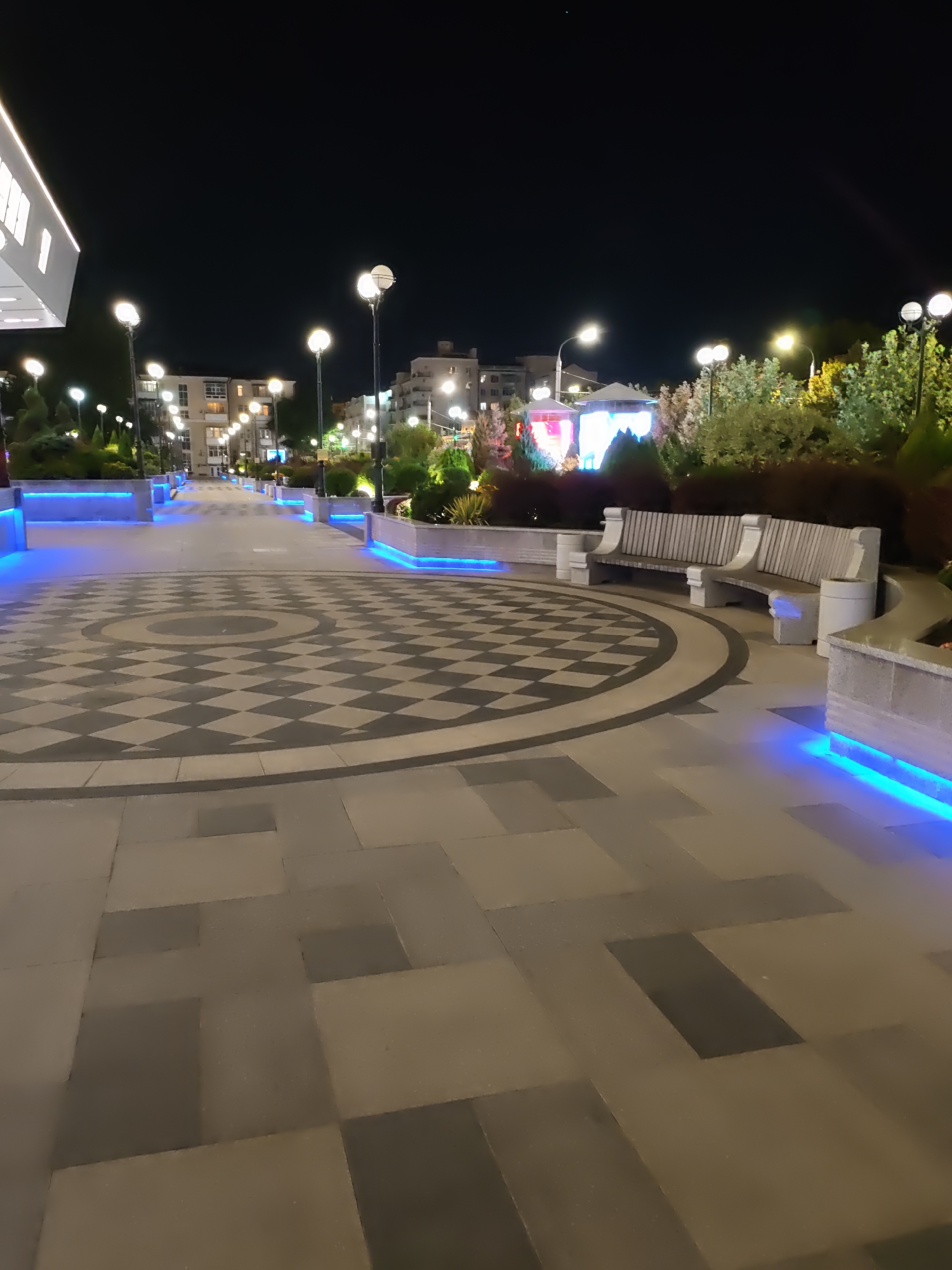 The image shows an outdoor public space at night, likely a park or pedestrian area, in Rostov-on-Don, Russia. The ground is paved with large rectangular tiles and features a large circular inlaid pattern of alternating dark and light grey diamonds. Blue LED lights are embedded along the edges of raised planters and pathways, illuminating the area.
In the foreground to the right, a grey stone bench with a slatted seat and back is positioned next to a cylindrical waste bin. Further into the scene, several streetlights with spherical bulbs illuminate a walkway lined with trees and shrubs. Buildings, likely apartment blocks, are visible in the background, with some windows lit. In the middle ground, to the left, part of a modern building with white cladding and illuminated windows is visible. The sky is dark, indicating nighttime. There are no people visible in the image.