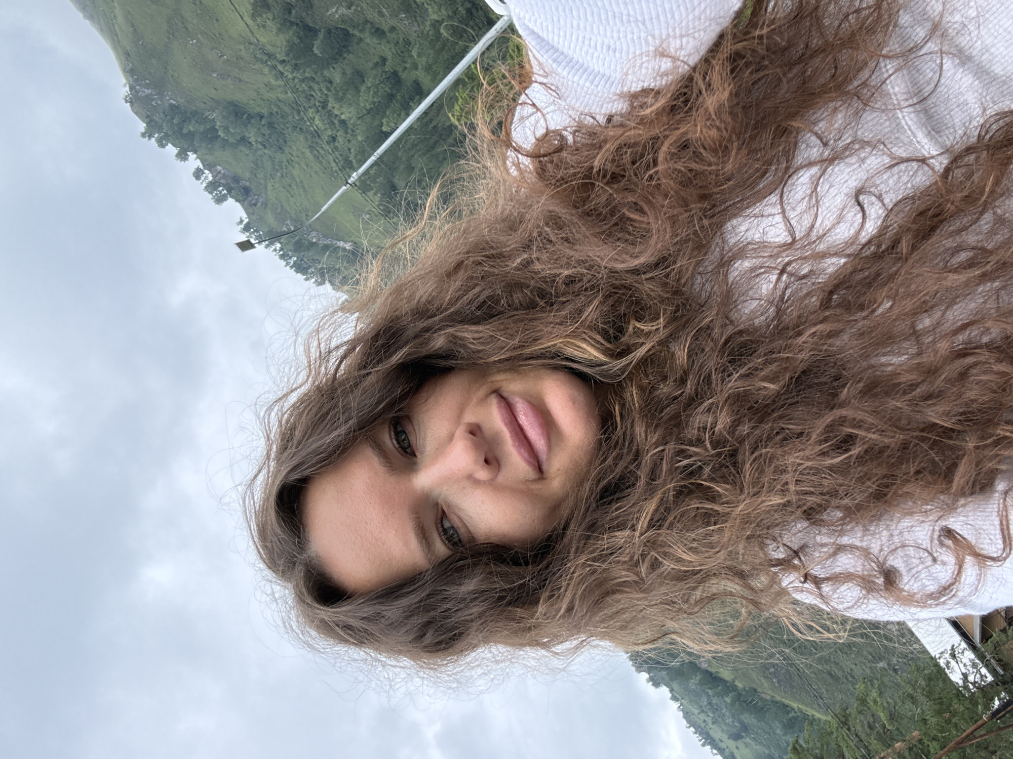 A woman with long, curly brown hair takes a selfie outdoors. She is wearing a white textured top. Behind her, a green, tree-covered mountain slopes upward under a cloudy sky. A tall, thin pole with some attached equipment is visible on the mountain. In the lower left corner, a portion of a wooden structure is visible. The location is Manzherok, Russia.
