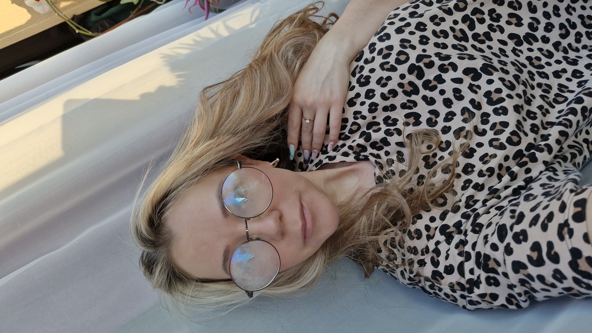 A woman with blonde hair and round glasses is lying down, wearing a leopard-print shirt. She has a ring on her finger and her nails are decorated. The location is unspecified.