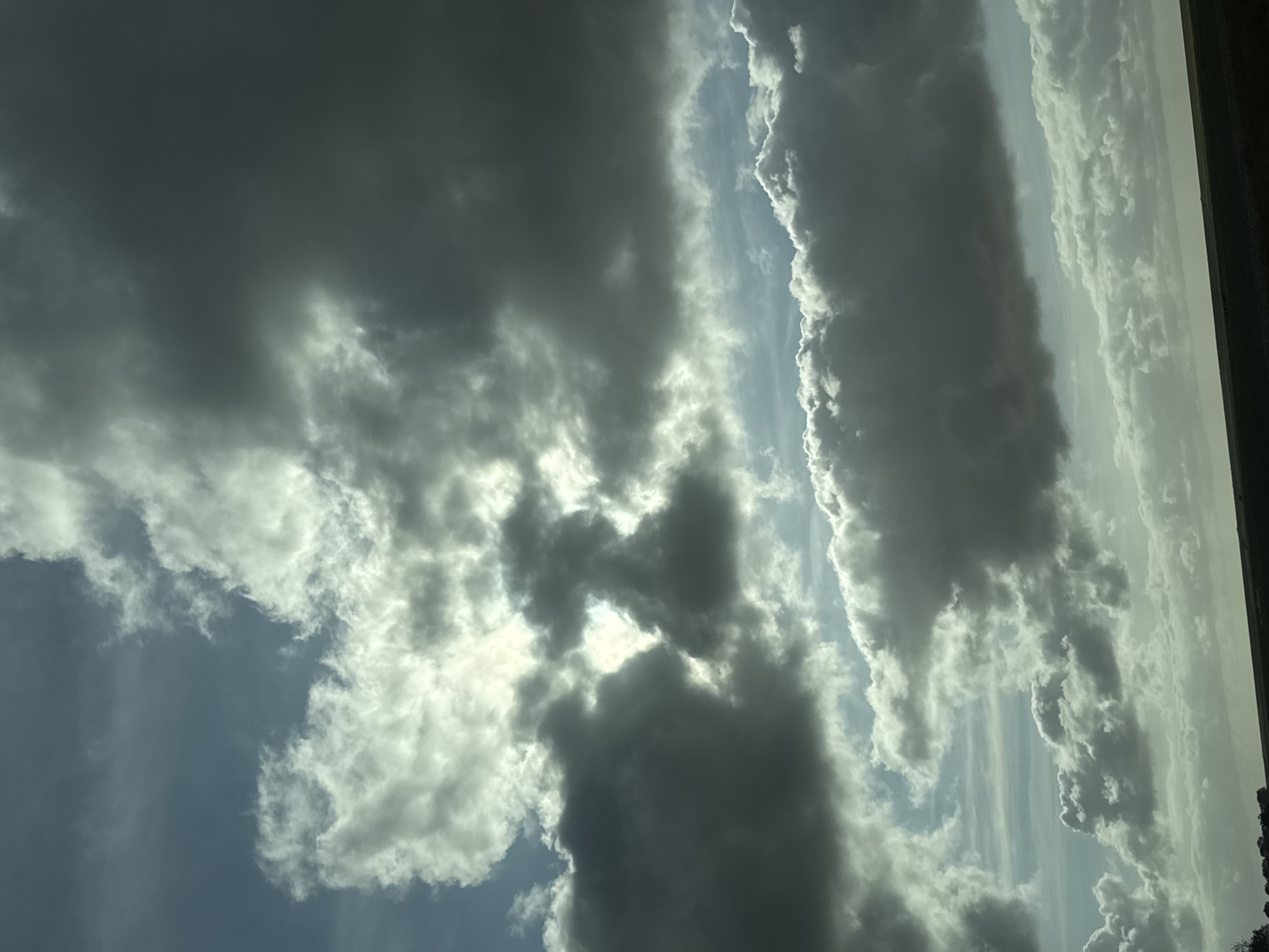 The image displays a sky dominated by a complex interplay of clouds under varied lighting conditions. The upper portion features dense, dark grey cloud masses interspersed with areas of bright, diffused sunlight shining through lighter cloud formations. A central bright opening in the clouds reveals an intense light source, casting strong illumination on surrounding cloud edges and creating high contrast with shadowed regions. Patches of pale blue sky are visible between the clouds, particularly towards the middle and lower left. Towards the bottom, a distinct horizontal line marks the horizon, beneath which a narrow, dark, indistinct strip of land or distant terrain is visible, appearing as a silhouette. No specific structures, people, or discernible objects are present other than the natural sky and a minimal suggestion of distant land.