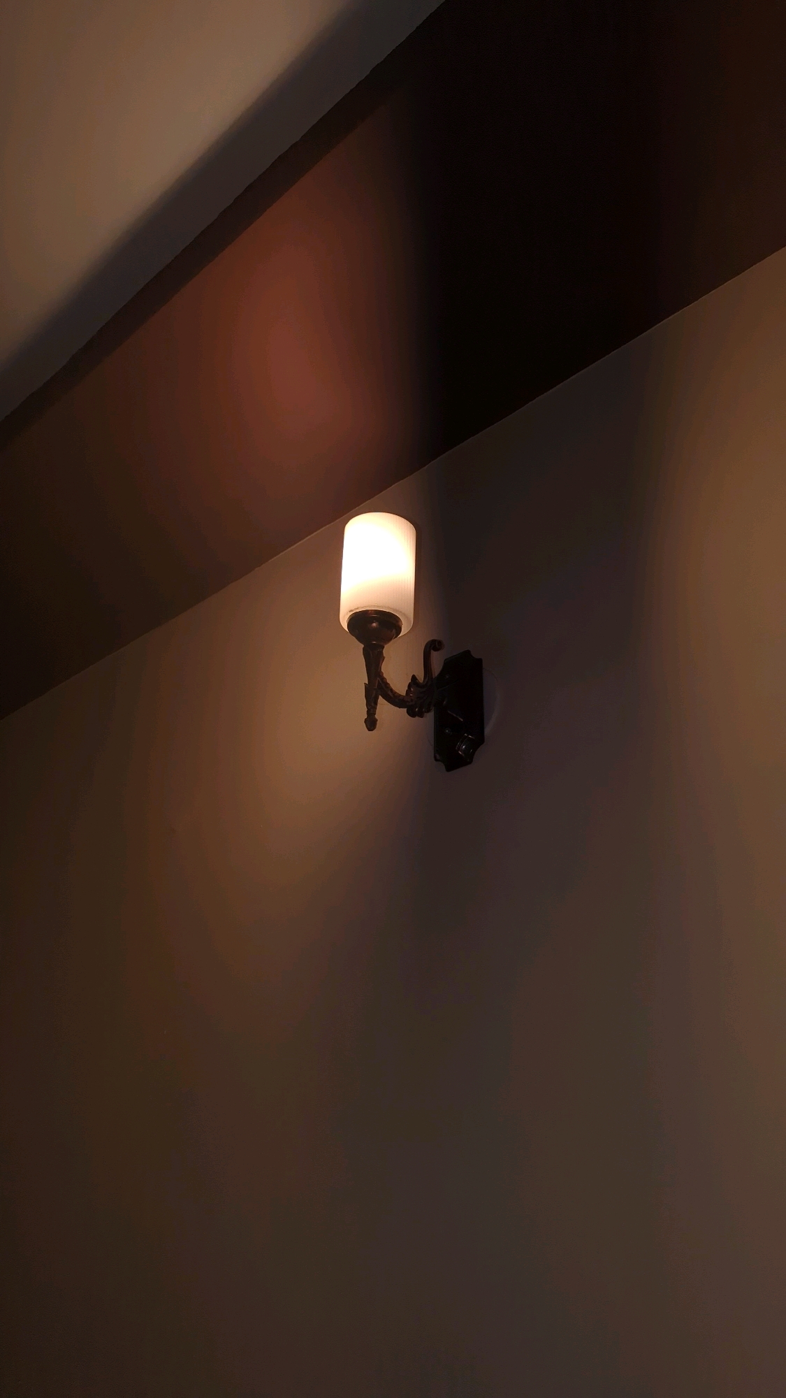A close-up, low-angle shot captures a single, lit wall sconce mounted on a muted, textured wall. The sconce features a dark, ornate metal base with a white, cylindrical glass shade emitting a warm, soft glow. The light casts a warm illumination onto the wall and ceiling, creating a gradient of light and shadow. The upper portion of the image reveals a dark, angled beam or architectural element, further defining the space. The overall impression is one of understated elegance in a dimly lit interior. The image lacks any discernible connection to a specific location within Karachi Division, Pakistan, as it focuses solely on the decorative lighting fixture and its immediate surroundings.