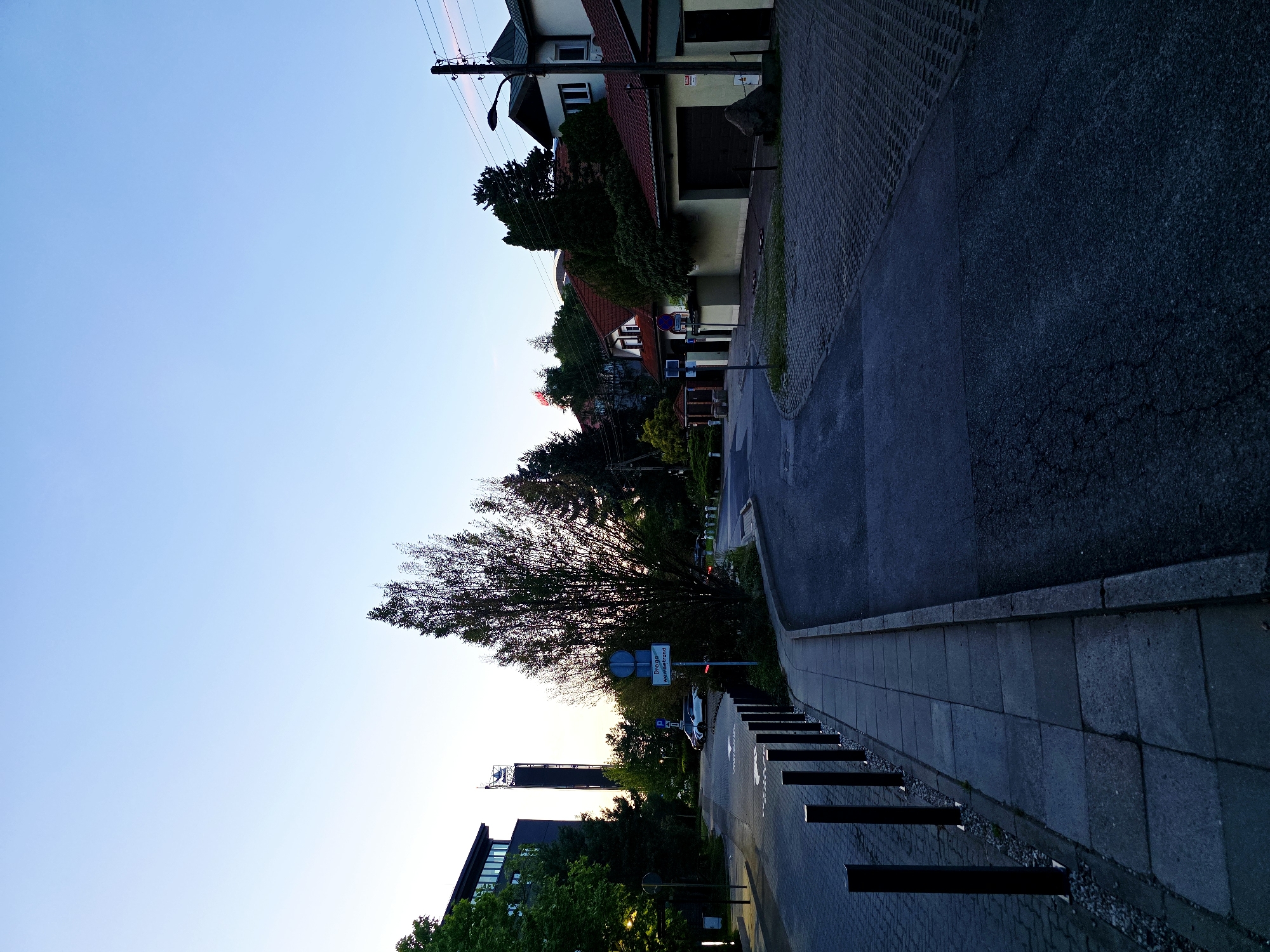 The image depicts a street scene in Warsaw, Poland, during the late evening or early morning, as indicated by the soft, diffused lighting and clear sky. The perspective is from a sidewalk looking down a curved asphalt road.
On the left side of the road, there is a paved sidewalk bordered by black bollards. Behind the sidewalk, a modern, dark-colored building with large windows is visible, partially obscured by trees. Further down the road on the left, a tall tower with a sign at the top, possibly a company logo or landmark, stands against the sky.
The road itself is asphalt, with some areas appearing darker and potentially wet. On the right side of the road, there are residential buildings with red-tiled roofs and light-colored walls. A prominent feature on the right is a wooden utility pole with wires and a streetlamp. A large rock is situated in front of one of the houses.
Several cars are parked or driving in the distance. A traffic sign is visible on the left, reading "Droga Wewnetrzna", which translates to "Internal Road" in Polish. The environment is characterized by well-maintained infrastructure, including paved surfaces and bollards, suggesting a developed urban area. The overall impression is a quiet, orderly street in a residential or mixed-use district of Warsaw.