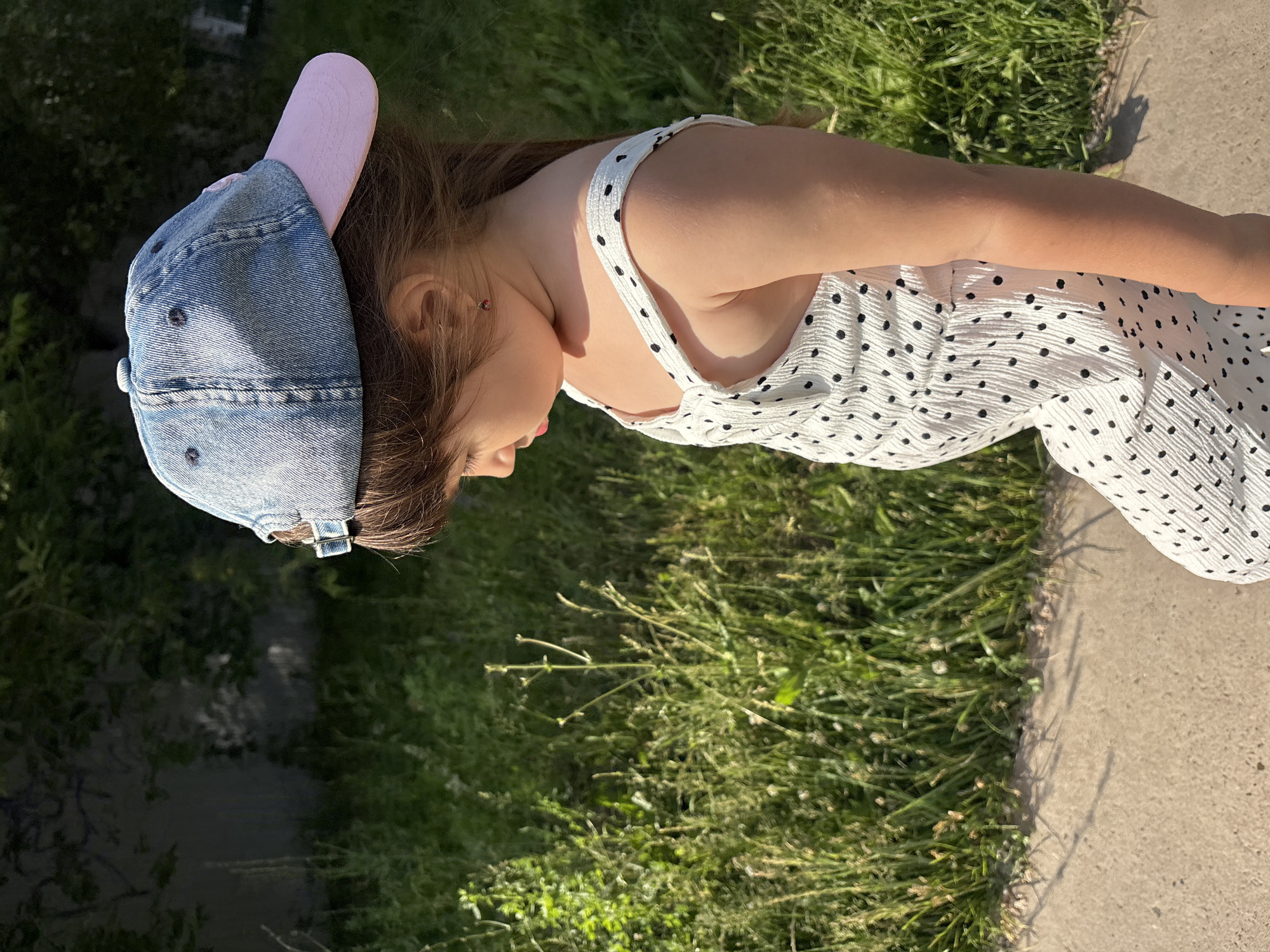 A young girl is captured in a close-up, sideways shot, leaning forward. She wears a denim baseball cap with a light pink brim and a white dress adorned with black polka dots. Her hair, a medium brown, is visible beneath the cap. The background consists of lush green grass and a concrete path, suggesting an outdoor setting. Sunlight casts shadows, indicating it is daytime, likely a sunny day. The girl's head is tilted downwards, and her facial expression is partially obscured, but there are no overt signs of distress or strong emotion. The image is oriented vertically, with the girl's body running from the top left to the bottom right of the frame. No text is visible in the image. The location context is Chaikovsky, Russia.