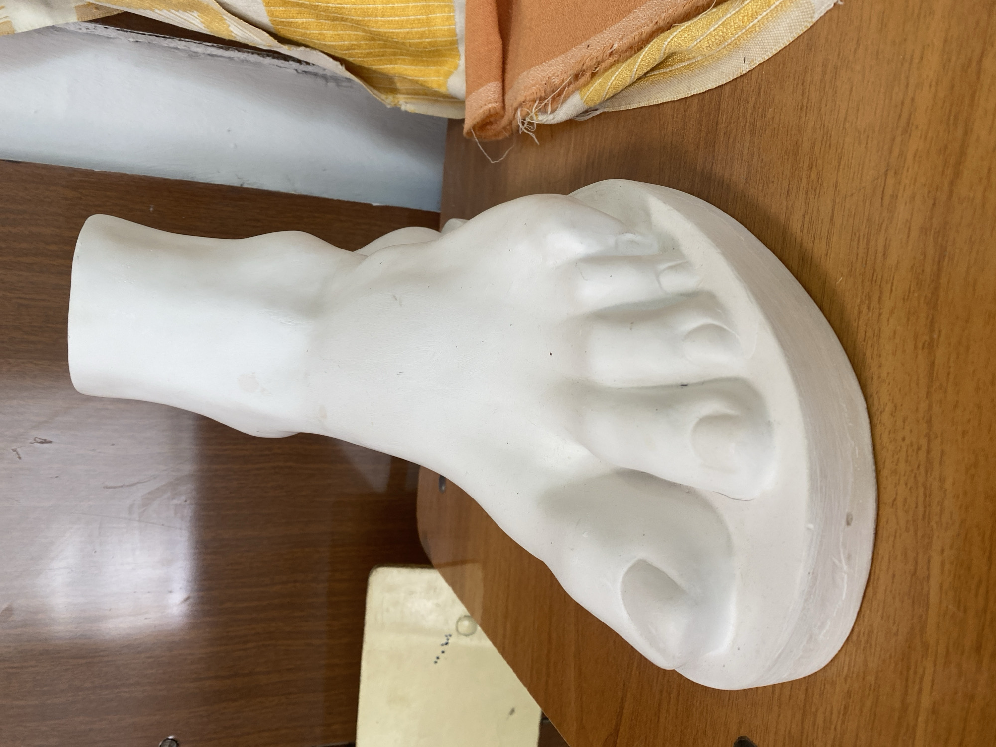 A white plaster cast of a foot rests on a wooden surface. A piece of fabric with orange and yellow stripes is behind it.