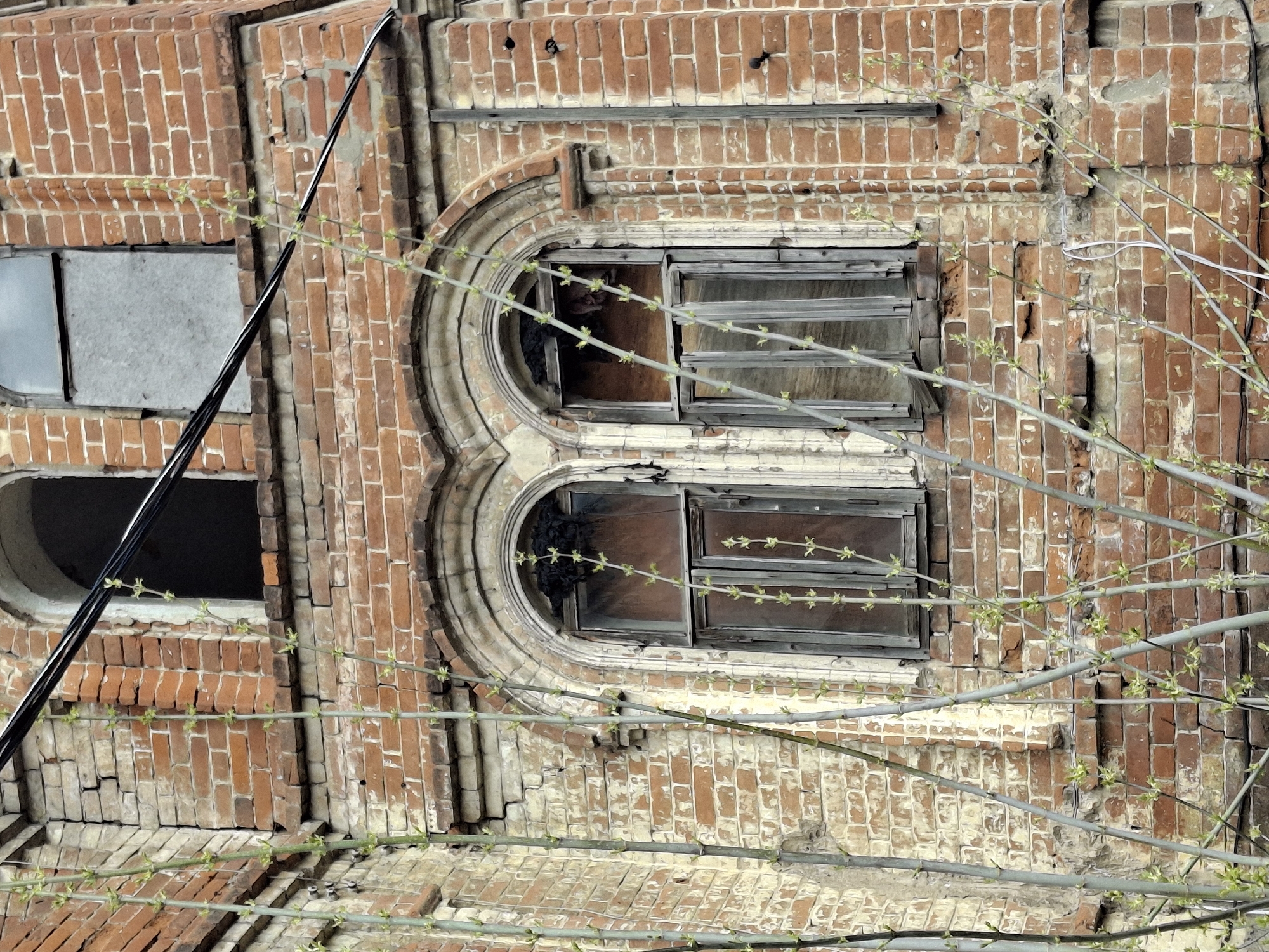The image shows a portion of a weathered, red brick building in Sarapul, Russia. Two arched windows, one above the other, are a prominent feature. The windows are boarded up, revealing damaged interiors. To the left, there are two other rectangular windows, one with visible gray covering and one hollow. Electrical wires are seen diagonally across the building's facade on the left side. Bare branches with sparse greenery partially obscure the building's front. The building shows signs of age and disrepair with crumbling bricks and faded paint.