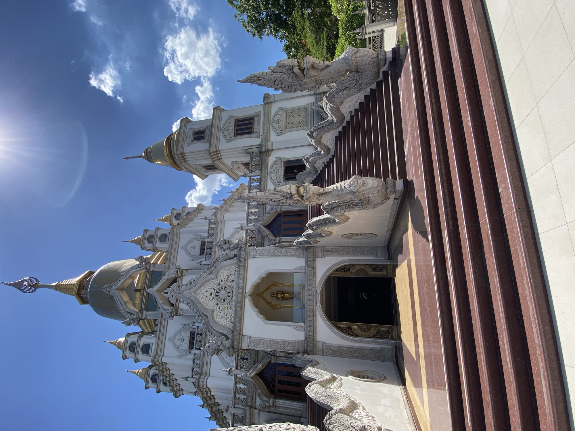 A low-angle, vertical shot of a white and gold temple with a brown stairway in Dĩ An, Vietnam. The temple is highly ornate with detailed carvings. A gold statue is visible through an archway. The staircase has serpent figures on either side. The sky is blue with white clouds. The sun is visible in the top left corner of the frame.