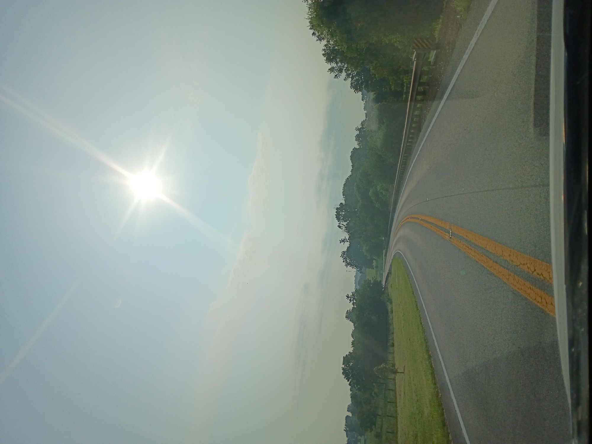 The image shows a bright, sunny day with a clear blue sky in Harrodsburg, United States of America. The sun is high in the sky, casting a strong glare. A curved road with double yellow lines runs diagonally across the frame, bordered by a grassy verge on the left and a dense line of trees on the right. The perspective suggests the photo was taken from a moving vehicle, possibly a car, looking out the window. The road appears to be an asphalt surface. The overall scene is serene and tranquil, with the natural landscape dominating the view. There is no visible text in the image. The time of day is likely midday due to the intense sunlight and clear sky. The weather is fair and sunny, with no clouds obscuring the sun. The location is identified as Harrodsburg, USA, which, combined with the rural scenery, suggests a drive through the countryside.