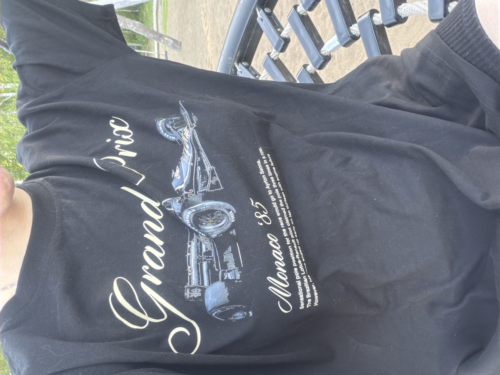 A black t-shirt with a graphic of a race car and the words "Grand Prix Monaco '85" is partially visible. The t-shirt is worn by a person, and the environment shows an outdoor setting with playground equipment in the background.