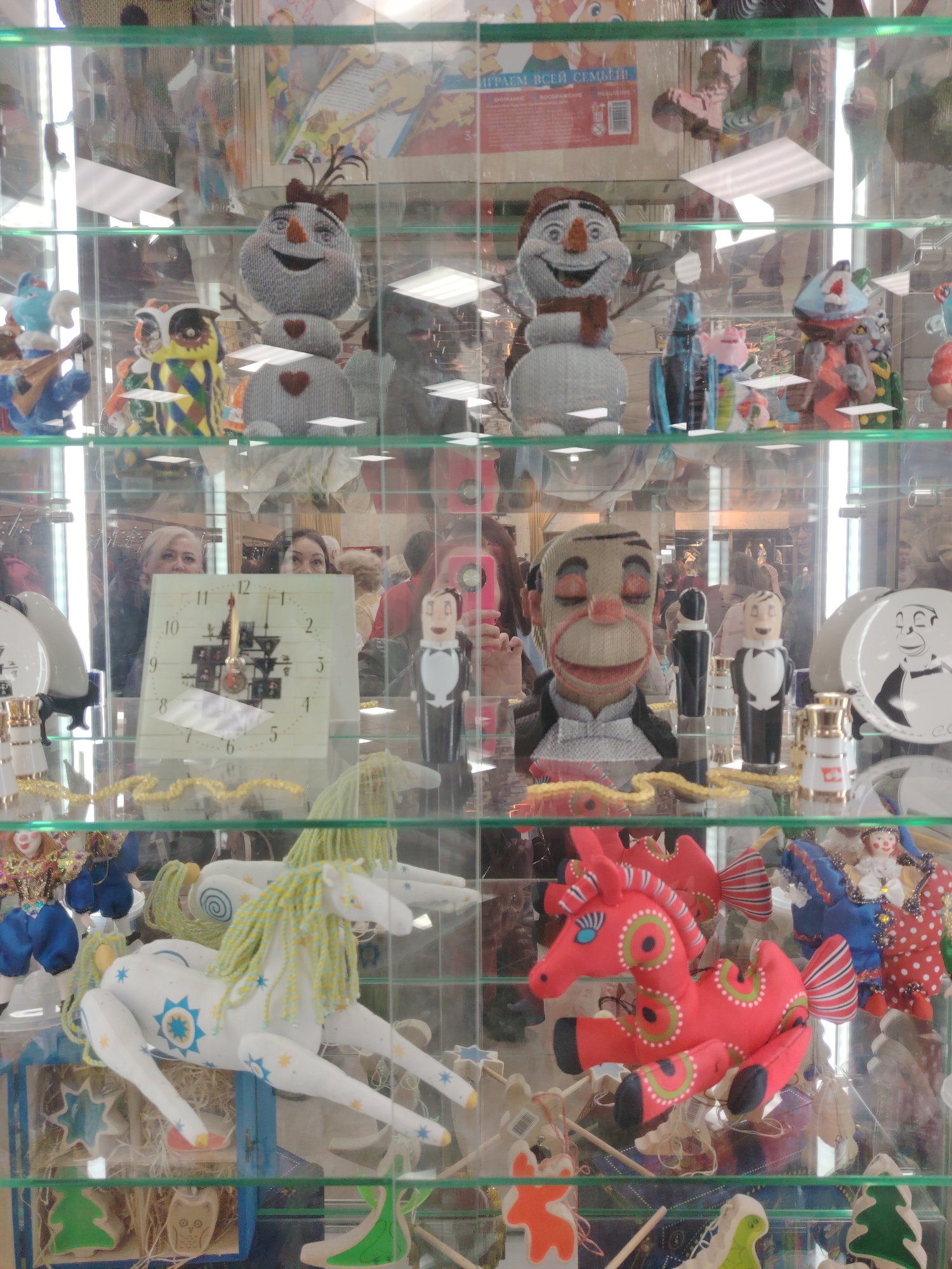 The image captures a display case, possibly in a shop in Moscow, Russia, filled with an assortment of folk art and crafts.
On the top shelf, two handmade snowman figures are displayed alongside painted ceramic figures.
The middle shelf features a clock with a square face, positioned alongside a puppet with a painted face and dressed in formal wear. Porcelain plates with painted faces and ceramic figurines stand nearby.
The bottom shelf is dominated by two stuffed horses, one white with a yellow mane and the other red with green detailing. Other handcrafted items, including wooden ornaments and figurines, are also present.
Reflections of people, including a woman holding a phone, are visible in the glass of the display case.