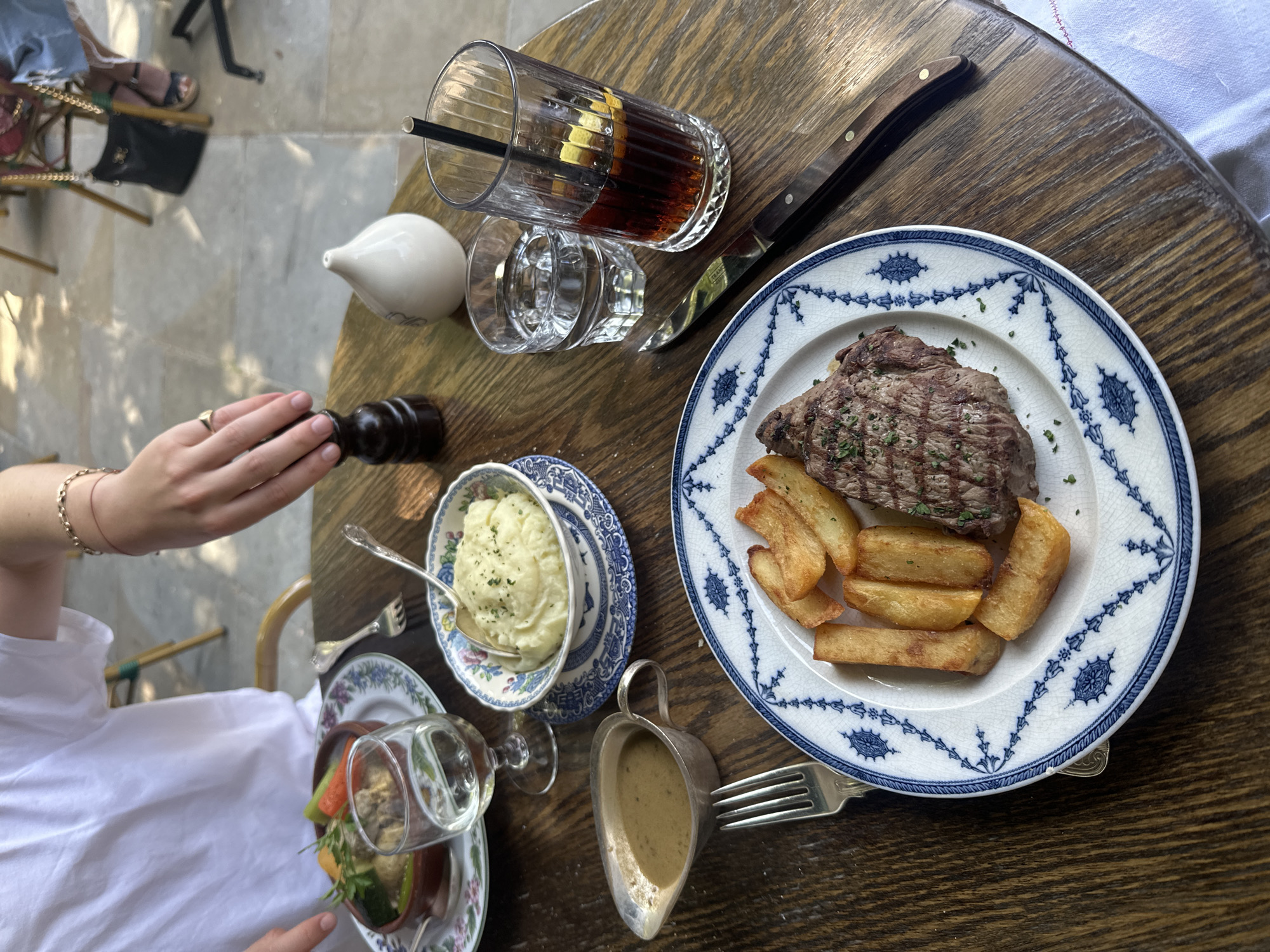 An overhead shot shows a wooden table at a restaurant. A diner with fair skin and a gold ring on their hand is holding a dark-brown pepper grinder. A plate of steak with large-cut fries is present. Beside the plate, there's a gravy boat and a fork. Adjacent to this is a bowl of mashed potatoes in a patterned bowl, along with a salad. Drinks include a cola with a lemon slice and a straw, a small white ceramic pitcher, and an additional glass. A knife also rests on the table. The setting appears to be an outdoor restaurant with other patrons visible in the background.