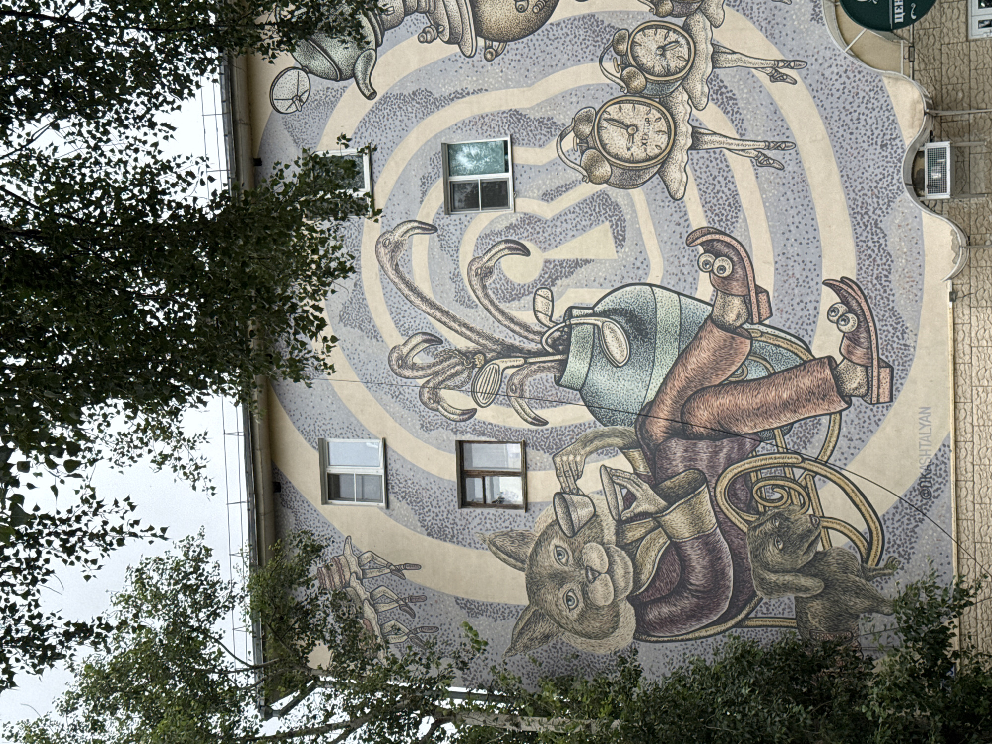 This media file is a photograph of a building mural in Nizhny Novgorod, Russia. The mural depicts a whimsical and surreal scene with a variety of anthropomorphic characters and fantastical elements, rendered in a detailed, illustrative style.
The central figures appear to be stylized animals and creatures, including what looks like a rabbit with large eyes, a furry, four-limbed creature, and disembodied hands and feet. These characters are engaged in what seems to be a playful or perhaps chaotic activity, interacting with various objects. Among these objects are multiple clocks with visible hands and Roman numerals, suggesting a theme of time or a surreal, dreamlike state. There are also elements resembling mechanical parts, tentacles or limbs, and a golf bag. The background of the mural is characterized by a swirling pattern of concentric circles in shades of gray and beige, interspersed with stippled textures.
The mural is painted on the exterior of a building, with several windows integrated into the artwork. Some windows appear to be open, reflecting the sky. The photograph is taken from a low angle, looking up at the building. Trees with dense foliage partially obscure the mural, with their branches and leaves framing the composition.
The visible text in the mural reads "@KASHTALYAN", which is likely an artist's signature or watermark. The presence of trees and the overcast sky suggest an outdoor setting, possibly on a cloudy day. The overall impression of the mural is one of surrealism, fantasy, and playful imagination.