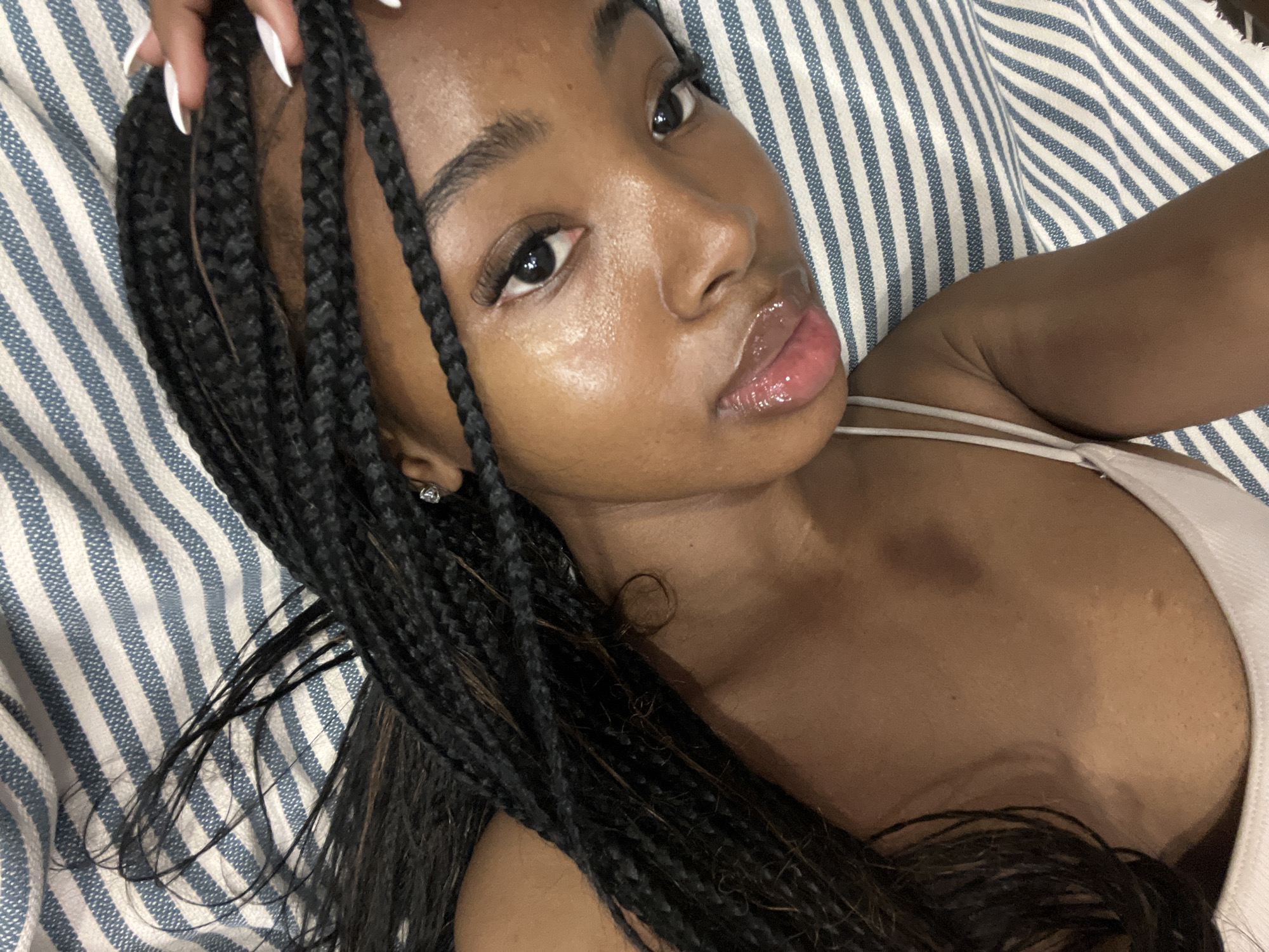The image shows a close-up, overhead view of a young Black woman with dark braided hair. She is lying down, with her head tilted back and her gaze directed upwards. Her eyes are open, and she is wearing light-colored makeup, including lip gloss. She is wearing a light-colored tank top. Her right hand, with white manicured nails, is resting on her hair near her forehead. The background consists of a striped fabric with alternating white and blue stripes. The image does not contain any information that would allow for the identification of a specific location in the city of Kazan, Russia.