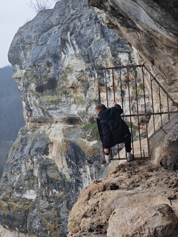 The image displays a person traversing a rusted metal gate positioned at a rock-cut entrance on the Shumen Plateau, near the city of Shumen, Bulgaria. The individual, appearing male, is clad in a dark winter jacket, dark trousers, and white sneakers. They are depicted with their left leg hooked over the top horizontal bar of the gate, while their hands grip the vertical bars, placing them in a precarious position over an outward drop. Their head is turned left, looking downwards. The gate, constructed from heavily rusted dark-brown metal bars, is angled into a rough, light-brown, eroded rock face that forms part of a cave or rock shelter entrance. To the left, a massive, multi-layered limestone cliff, mottled with gray, white, and green lichen, descends sharply. Sparse, bare tree branches are visible at the cliff's summit, and a distant, hazy landscape of hills or forests lies beyond the cliff face, under an overcast sky.