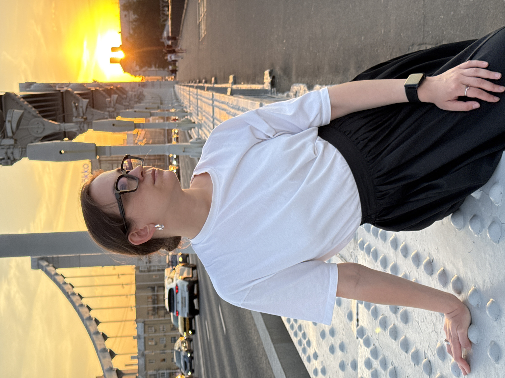The image shows a woman lying on the railing of the Krymsky Bridge in Moscow, Russia, during sunset. She is wearing glasses, a white t-shirt, a black skirt, and a wristwatch. Her left hand rests on the railing. In the background, traffic flows across the bridge, and the sun sets behind buildings. The bridge's distinctive metal structure is visible.