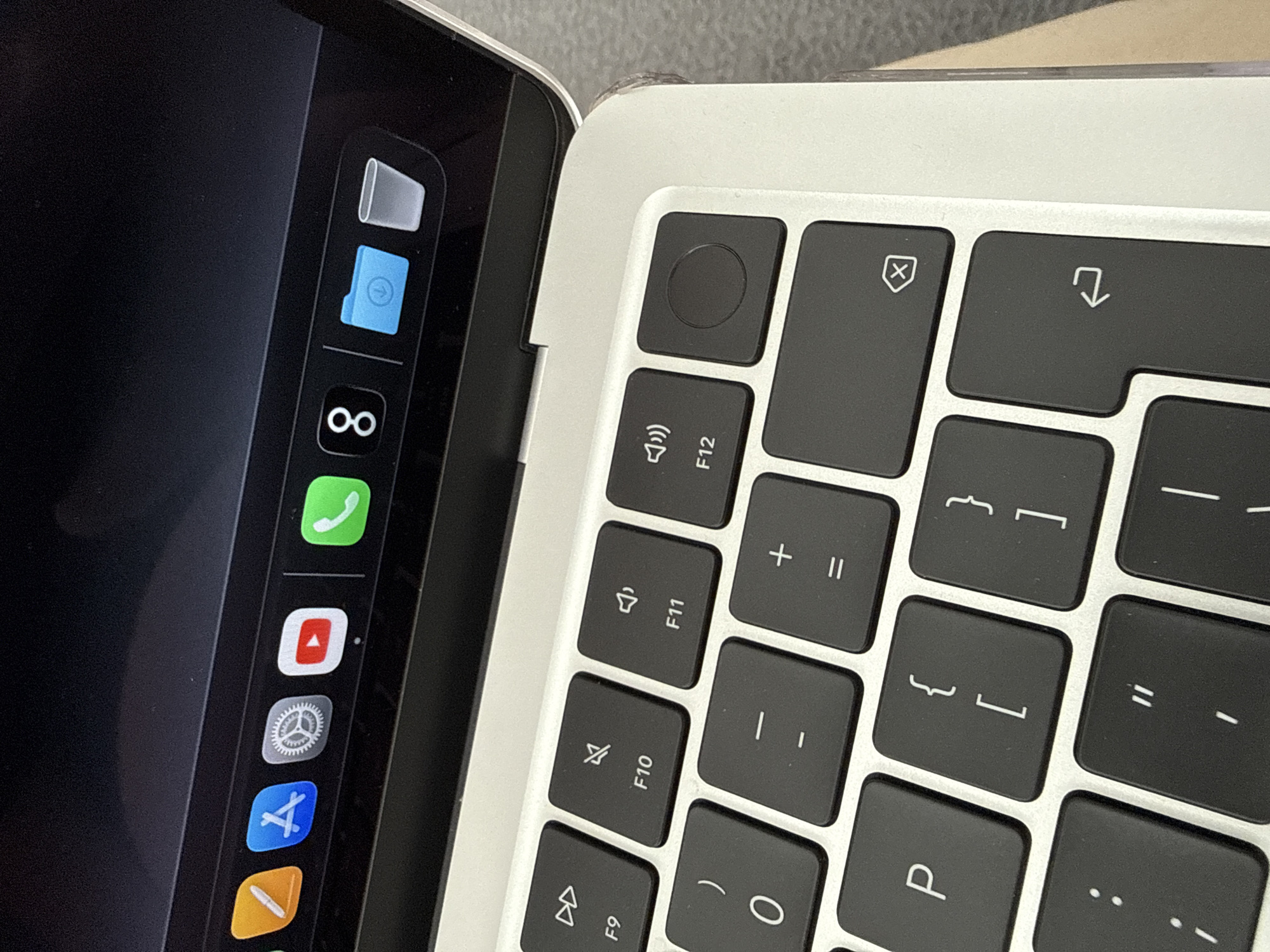 A close-up shot depicts part of a laptop with the dock visible on the screen and a section of the keyboard showing. The dock on the screen includes icons for the trash, a folder, an infinity symbol, a phone, YouTube, settings, and other applications. The keyboard shows the upper right portion, including keys like escape, volume, brightness, and function keys F9 through F12, as well as the characters '{', '}', 'P', and the shift key. The keyboard and screen are silver in color with black keys. The setting and geographic location are not evident in the image.