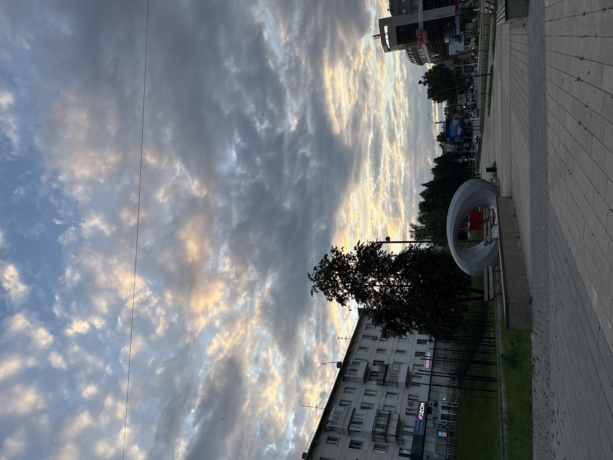 The image depicts a street scene in Chaikovsky, Russia, at dusk. The sky is filled with dramatic, textured clouds, tinged with the warm hues of sunset. In the foreground, a paved pedestrian walkway with square tiles leads towards a street. To the left, a multi-story apartment building with numerous windows and balconies is visible, with a grassy slope leading up to it. A dark green tree stands prominently in front of the building, partially obscuring the lower floors. In the mid-ground, a large, grey, modern art sculpture with an oval opening is situated near a concrete bench. Further down the street, a modern building with the "DNS" logo is visible, along with trees lining the road. Traffic lights and street lamps indicate the presence of a functioning urban environment. The overall impression is a tranquil evening in a city setting.