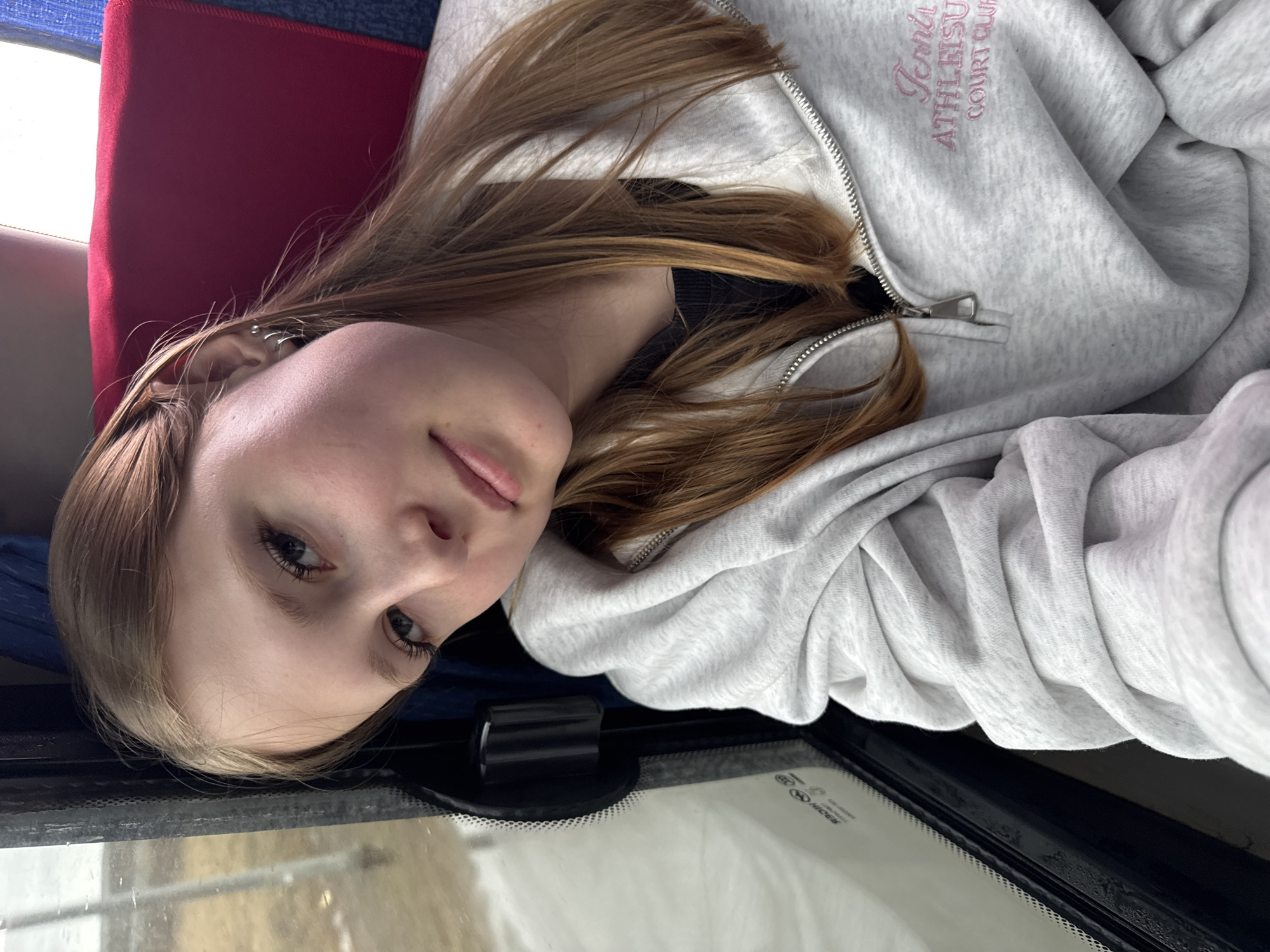 A young woman with light brown hair is positioned in what appears to be the interior of a vehicle or bus, evident by the seat and the window at the bottom of the image. She is wearing a light gray zippered hoodie with pink embroidery that reads "Tennis Athletics Court Club". Her head is tilted, and she looks directly at the camera. The vehicle's interior shows red and blue colored seating. The location is not discernible from the image, nor is there evidence provided that would place it specifically in Kukushtan, Russia.