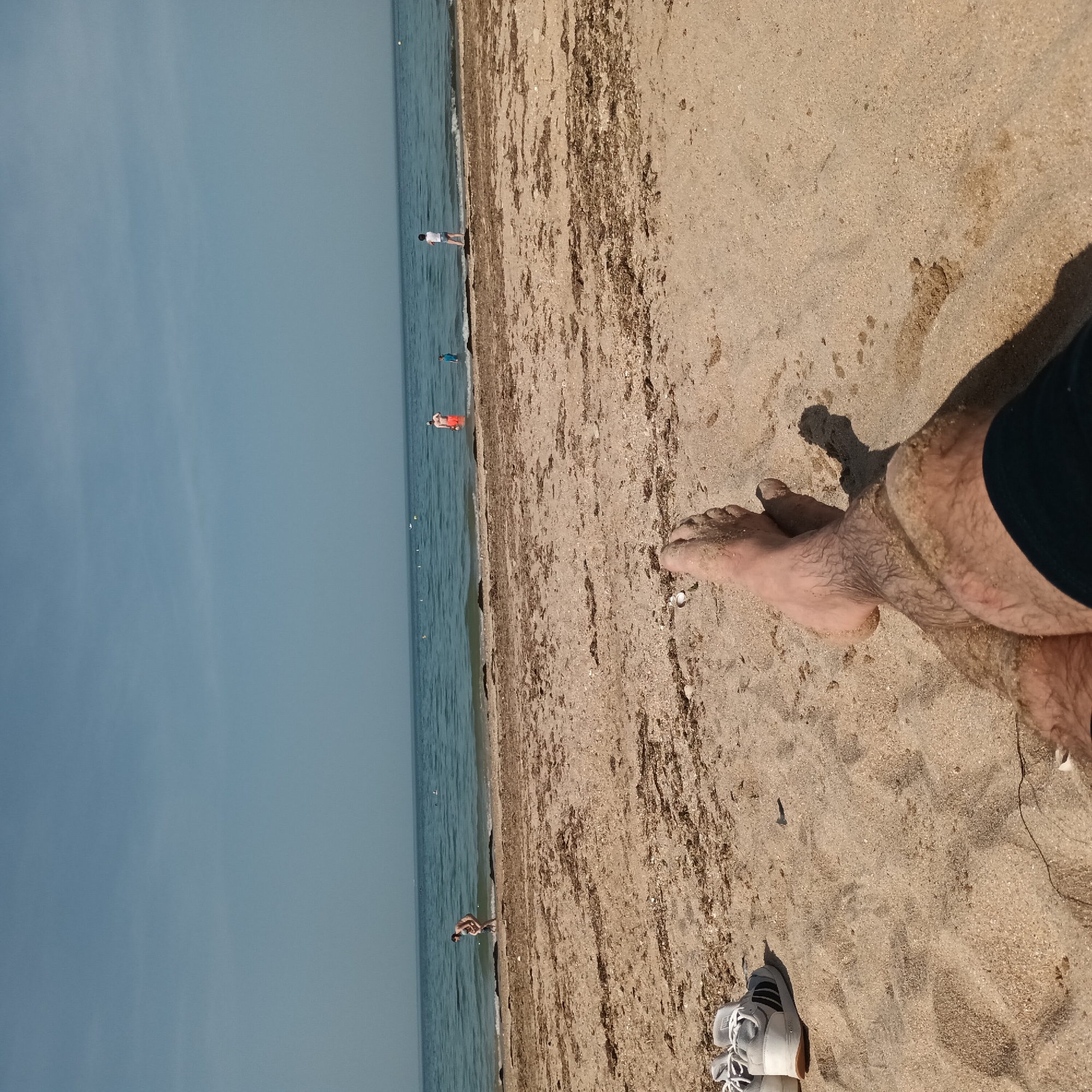 The image shows a beach scene. A light blue sky meets the darker blue sea, which is separated by a sandy beach covered with seaweed. Several people are in the distance, either in the water or along the shoreline. In the foreground, there are human legs and feet covered in sand, and a white shoe is lying on the beach. The location is unknown.