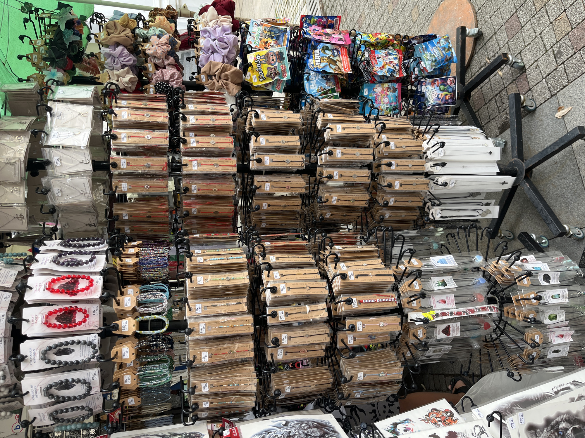The image depicts a street vendor's stall, likely in Sliven, Bulgaria, displaying a variety of accessories for sale. The stall is a multi-tiered, rotating rack filled with numerous small, packaged items. These include a wide assortment of bracelets and necklaces, many of which are beaded, made of stone, or feature small charms. Some items are displayed with price tags, indicating individual sales.
Prominently featured are sections dedicated to bracelets, some with dark grey beads (labeled "OBSIDIAN" and "GRANAT"), others with red beads, and many with colorful beaded designs. There are also numerous red string bracelets with small charms. Necklaces are displayed in clear plastic packaging, with various pendants such as hearts, crosses, and geometric shapes.
Towards the right side of the stall, a selection of hair scrunchies in various pastel and neutral colors are visible. Below the accessories, several packaged children's toys or collectible items are displayed, featuring characters from popular animated movies like "Inside Out" and "Paw Patrol."
The stall is set up outdoors, with a green awning overhead providing shade. The ground is paved with cobblestones, and a few fallen leaves are visible on the ground. The overall scene suggests a bustling marketplace or sidewalk sales environment. No individuals are clearly identifiable in the immediate foreground.