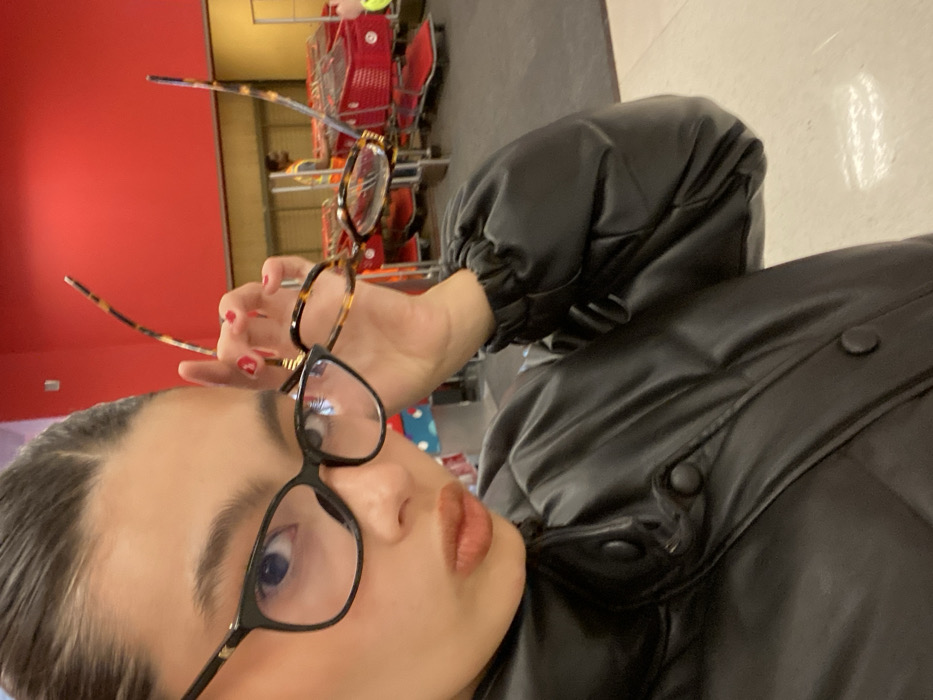 A person in a black puffer jacket holds two pairs of glasses up to their face, one with black frames and the other tortoiseshell. They are positioned in front of a red wall, shopping carts, and a grey floor, likely inside a store. The person has dark hair and is looking at the camera.