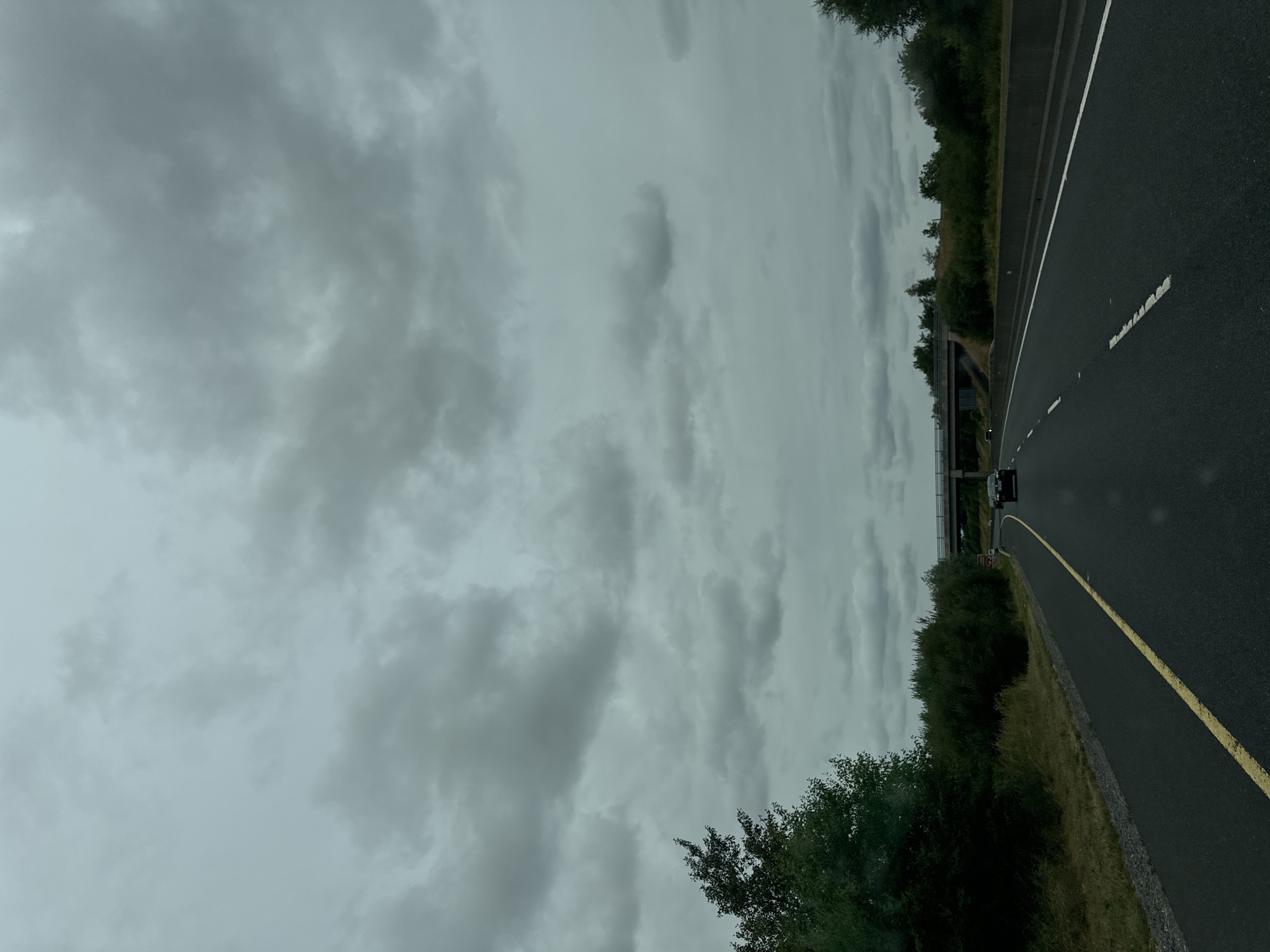 The image captures a view from a vehicle on a highway in Ireland. The sky is overcast with dense, grey clouds, suggesting a potentially rainy or gloomy day. The highway itself is paved and marked with a yellow line on the left and white dashed lines on the right. Vegetation, including trees and bushes, lines the sides of the road. In the distance, a concrete overpass or bridge spans across the highway. A dark-colored car is visible on the highway ahead, traveling in the same direction as the viewer. The scene is devoid of any people or significant landmarks to pinpoint a specific city or location within Ireland.