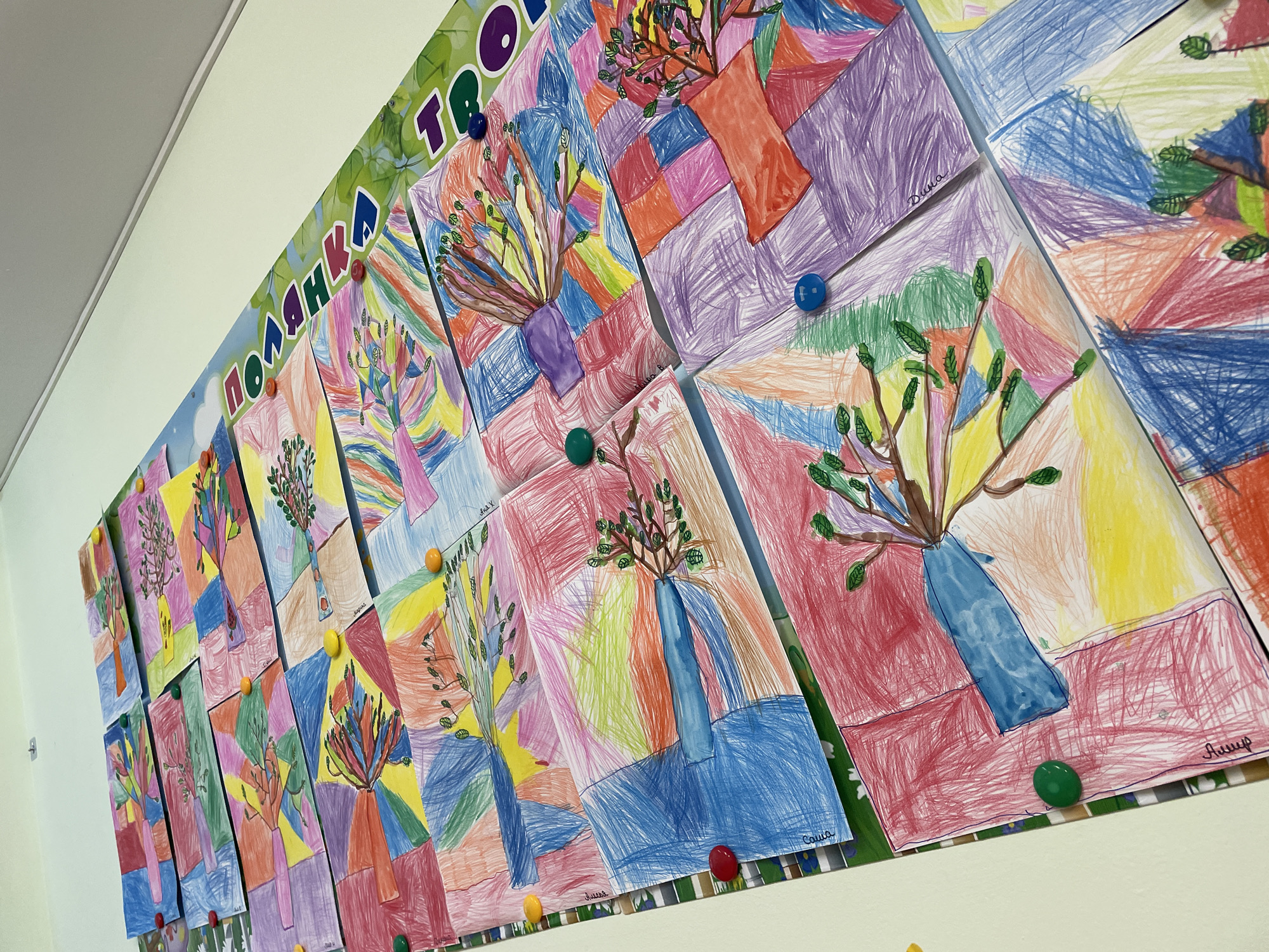 The image shows a display of children's artwork pinned to a wall under a sign with the word "ПОЛЯНКА ТВОР" (Polianka Tvor) in large colorful Cyrillic letters. The artwork consists of drawings of vases with branches, rendered in various colors and styles. Each drawing appears to be on a separate piece of paper, and the papers are attached to the wall with colored pushpins. The scene is likely indoors, possibly in a school or art center. While there is no information indicating the image was taken in Izhevsk, Russia, the Cyrillic lettering of the sign suggests a location where Russian is spoken.