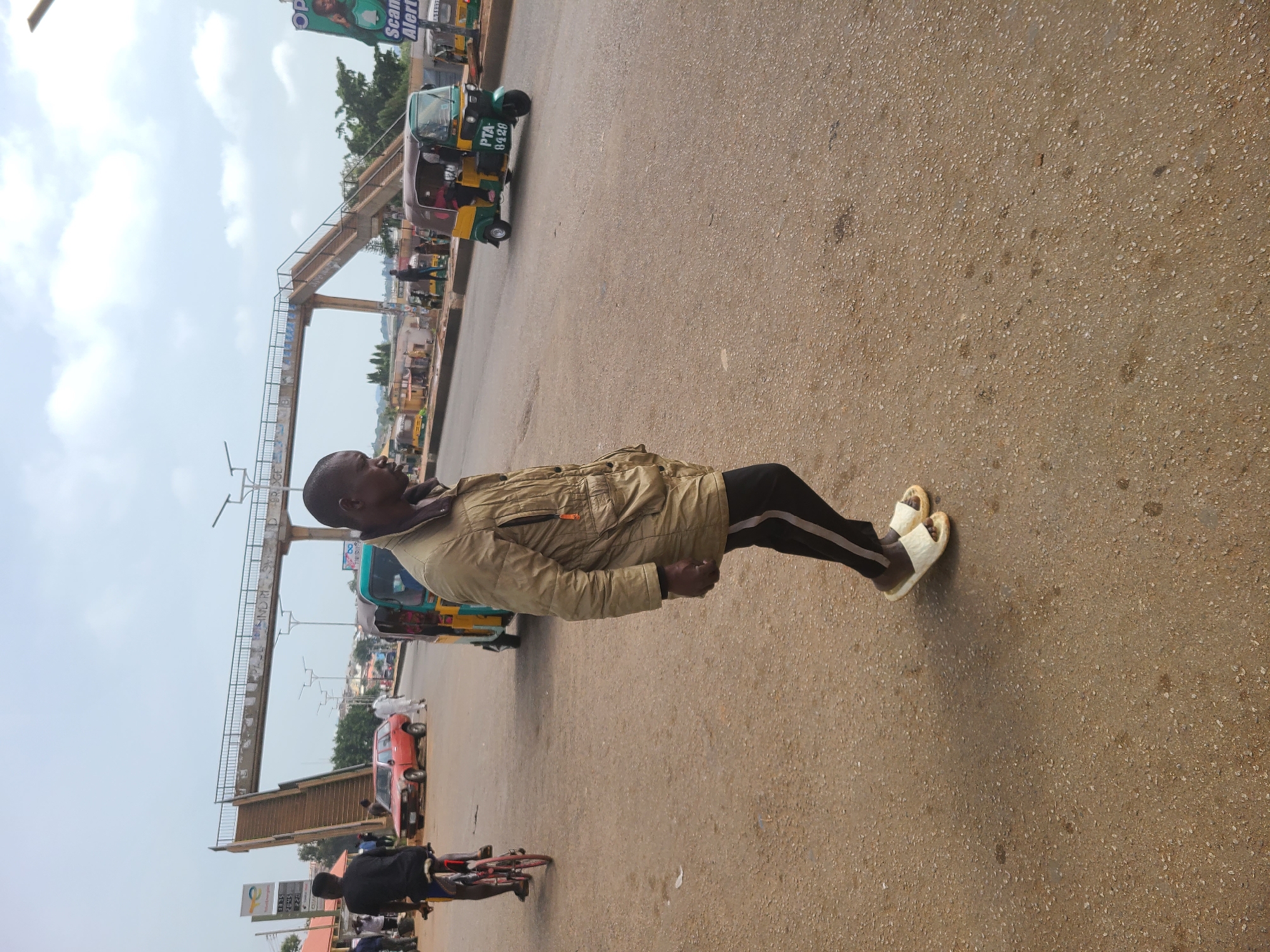 A man in a light brown jacket and dark pants stands on a light brown paved surface in what appears to be a city street. He is wearing cream-colored sandals. In the background, there are auto rickshaws, cars, and a pedestrian overpass. A person on a bicycle is visible in the lower left corner. The image appears to be taken at street level. The sky is overcast. The location is Unknown, Unknown.