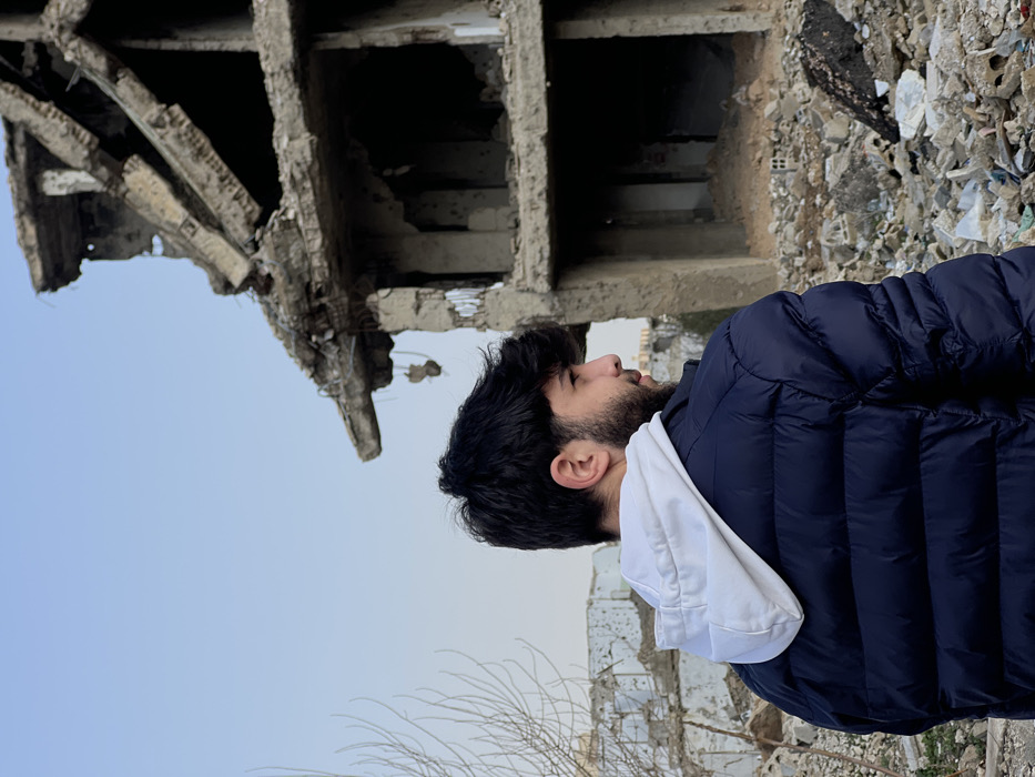 A young male, viewed from the upper back and right profile, stands amidst urban destruction in Joubar Municipality, Syria. He has dark hair and a beard, wearing a dark blue puffer jacket over a white hooded sweatshirt. His gaze is directed to the right of the frame. In the immediate background to the right, a multi-story concrete building stands severely damaged, with partial structural collapse, exposed reinforcement, and significant debris accumulated at its base. Further in the background to the left, additional heavily damaged concrete structures are visible. The ground throughout the scene is covered with extensive rubble, concrete fragments, and debris. Bare tree branches are present in the foreground on the left. The sky is clear and light blue.