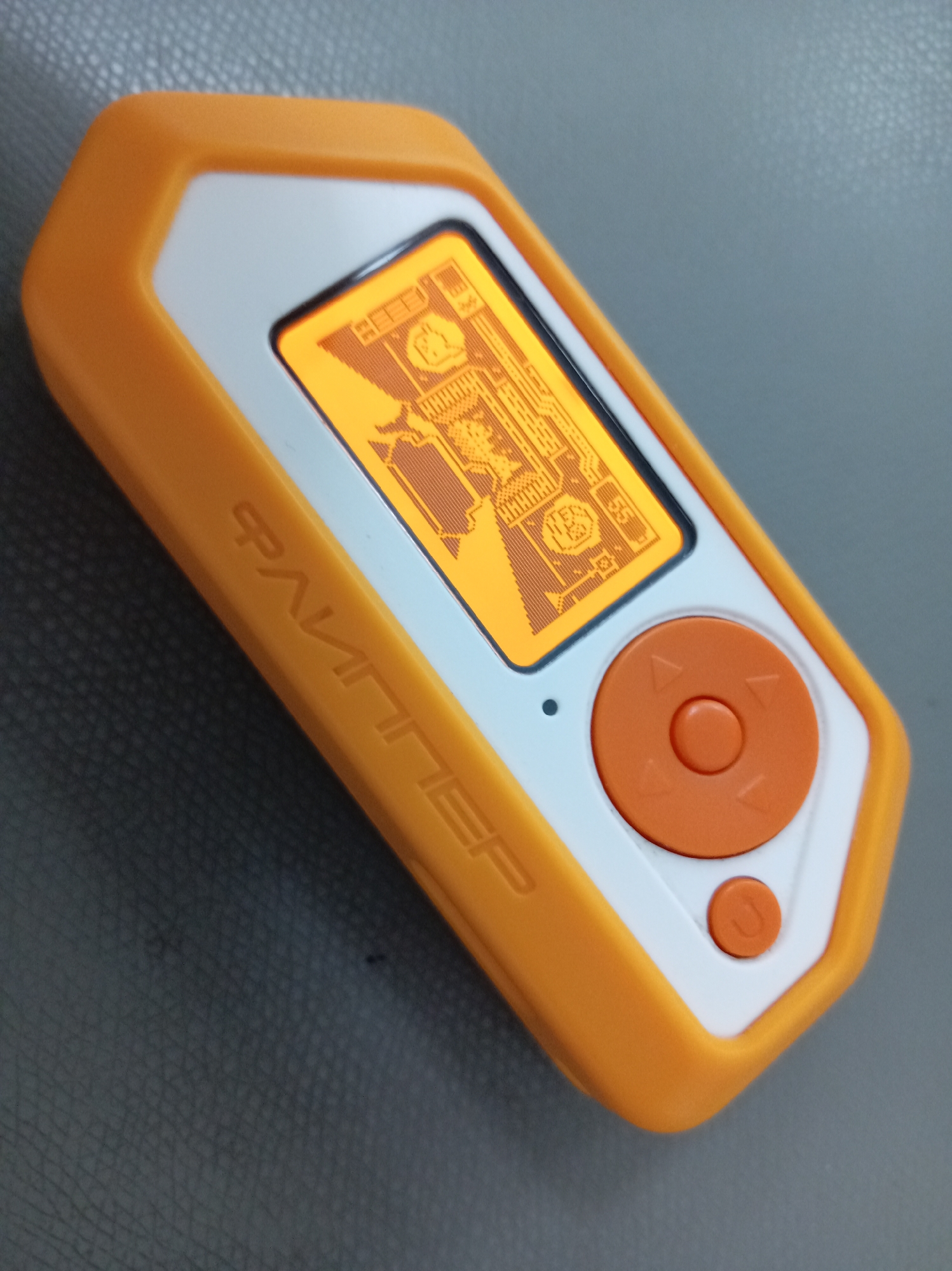An orange and white handheld device is shown from a high angle, resting on a dark gray textured surface. The device has a bright orange rubberized casing with a white inner bezel surrounding a rectangular screen. The screen displays a retro-style graphic in orange pixels, featuring what appears to be a schematic or game interface with various symbols and text. To the right of the screen is a circular orange button with directional arrows and a central button. Below this is a smaller, circular orange button. On the left side of the orange casing, the word "ФАЛЛИПЕР" is embossed in capital letters. The background surface has a subtle, repeating pattern, resembling leather or a similar material.