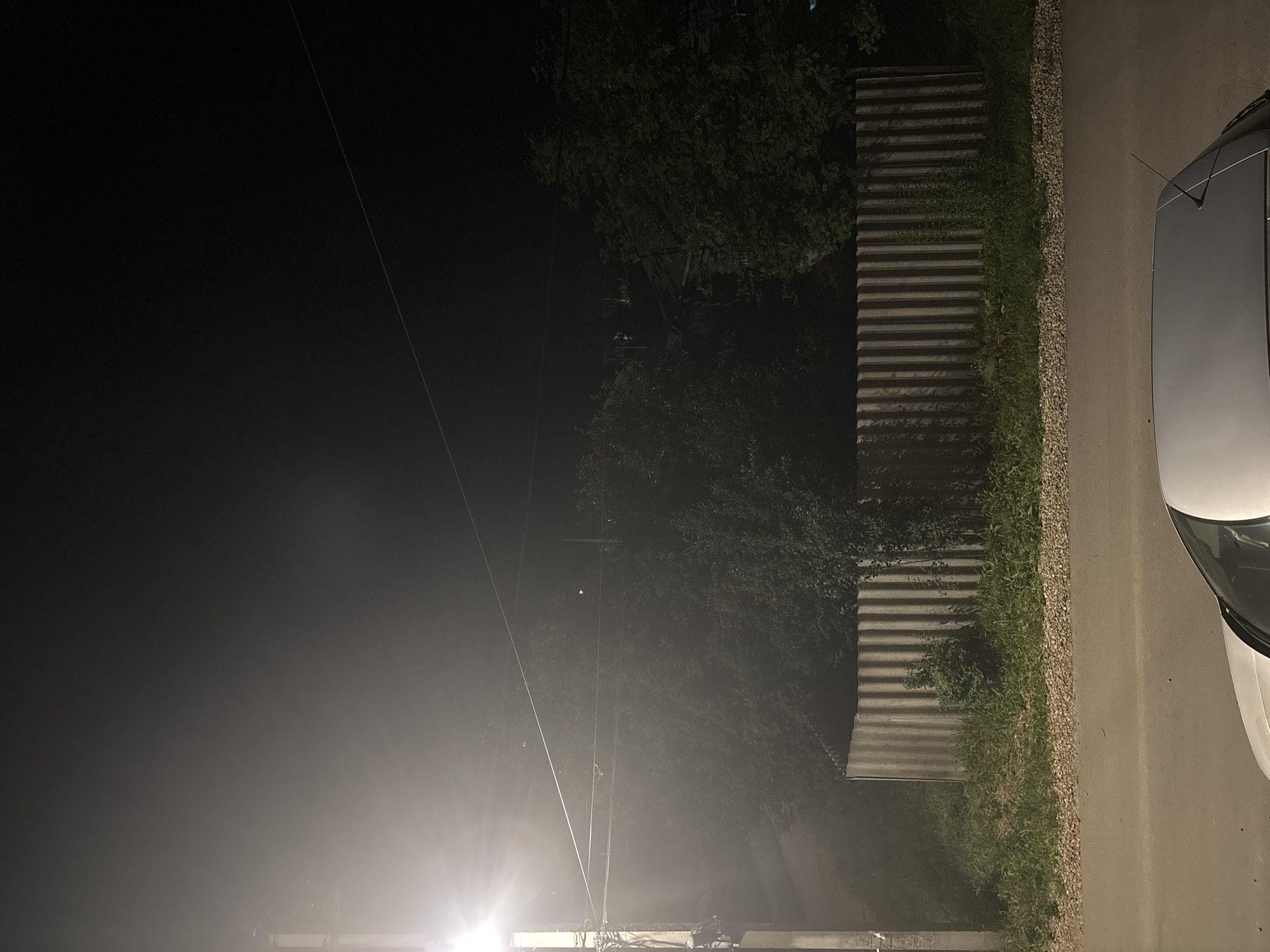 The image depicts a street scene at night in Kstovo, Russia. A streetlamp on the left illuminates a tall, segmented pole with wires attached. Beyond the pole, dense trees and foliage are visible, obscured by the streetlamp's glare and atmospheric haze. A corrugated metal fence runs horizontally across the middle ground, separating the trees from a patch of grass and gravel. In the foreground, the roof and antenna of a white car are visible, parked on an asphalt road. The overall environment suggests a residential or suburban area.