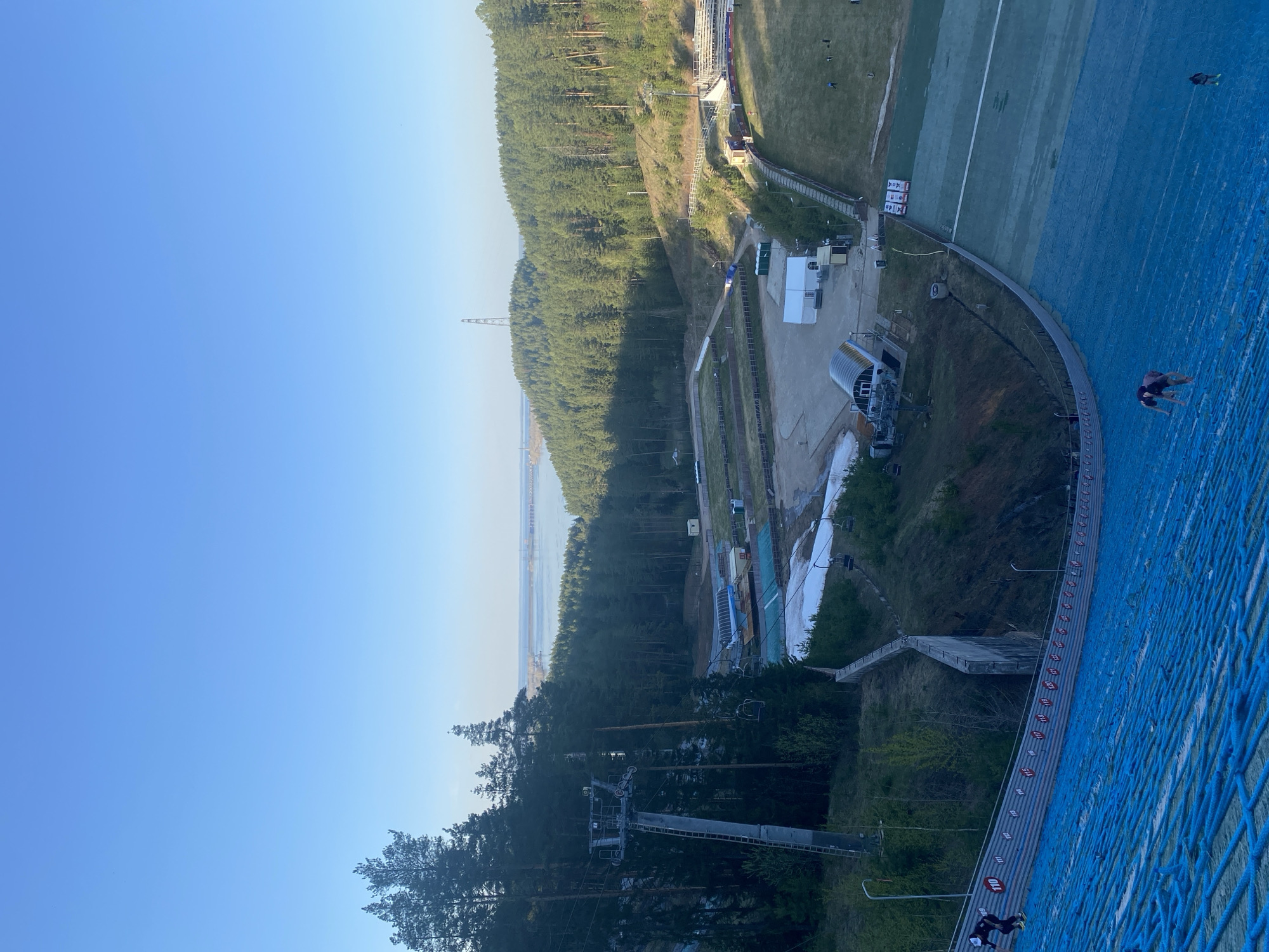 This image captures a view from the top of a ski jump in Прикамский, Russia. The jump is covered in blue netting, with a ladder running down the side marked with distance indicators in meters. A pair of individuals can be seen descending the ladder. The landscape includes a dense forest stretching toward a body of water with a bridge or causeway visible in the distance. A ski lift is partially visible amidst the trees. Below the ski jump, there is an area that includes buildings, snow-making equipment, and a grassy landing area. The sky is clear and blue.